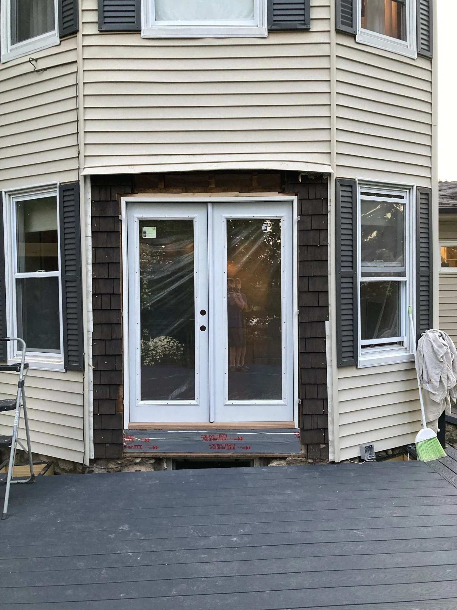 Back of a house with a newly installed glass sliding door, surrounded by beige vinyl siding and dark brown wood shingles. There are double-hung windows with shutters on either side of the door, with sunlight reflecting off the glass.