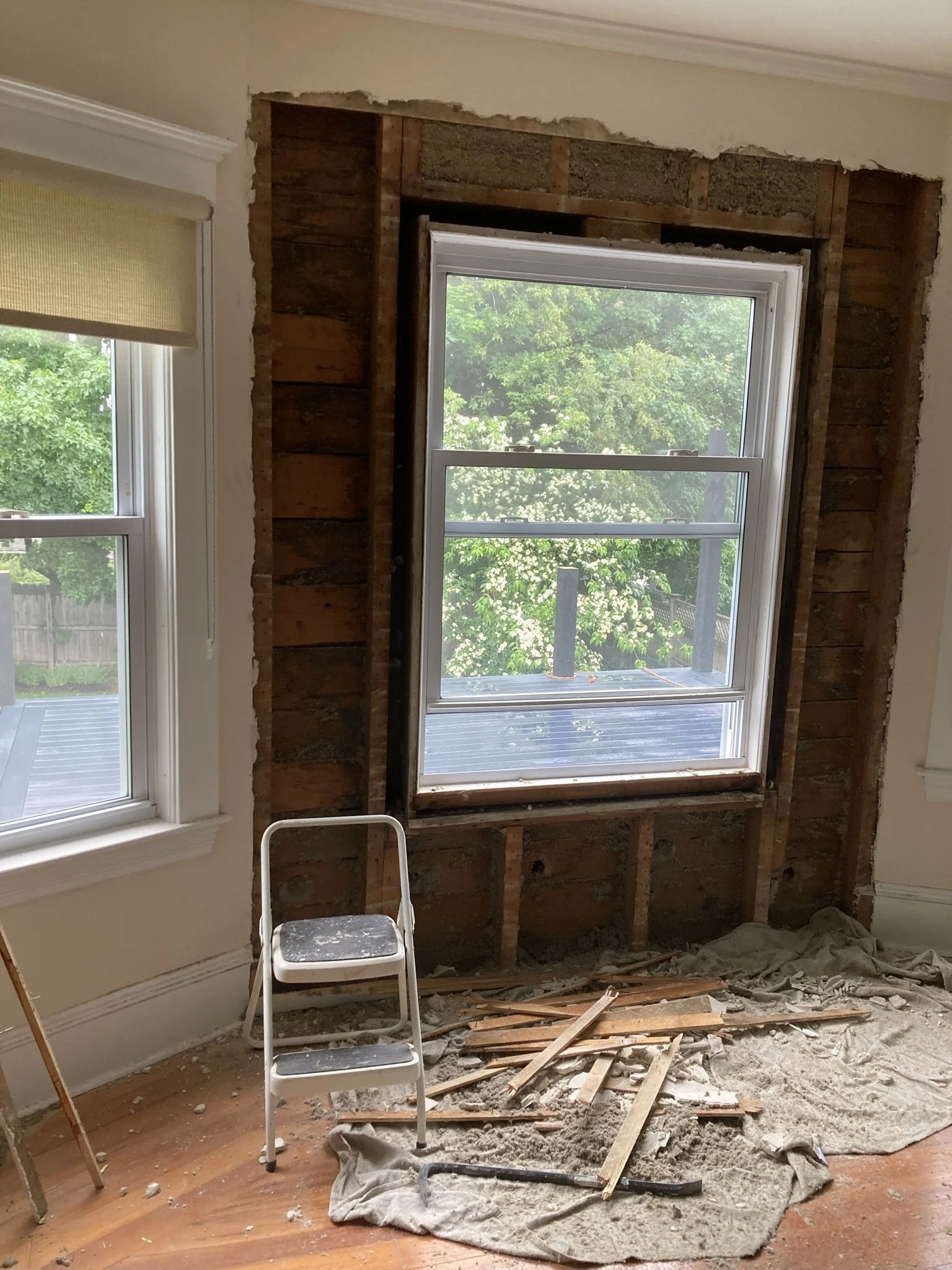 Room undergoing window replacement or renovation. Part of the wall around the window is removed, exposing the wooden framing and insulation. The window frame is intact, and outside greenery is visible. Construction debris and tools are on the floor, 