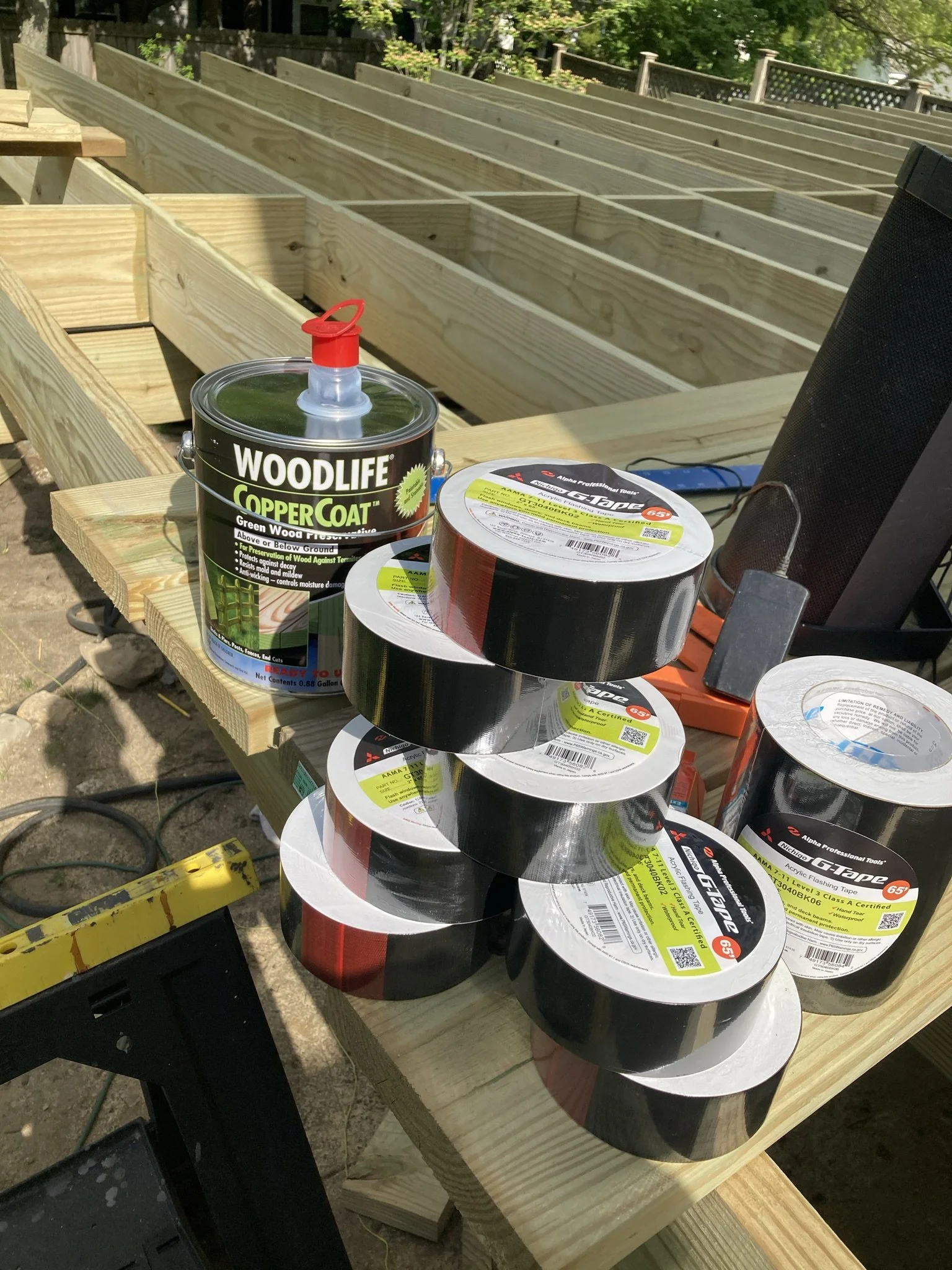 Stack of rolls of black adhesive tape, a can of wood preservative, and a black speaker on a wooden workbench outdoors.