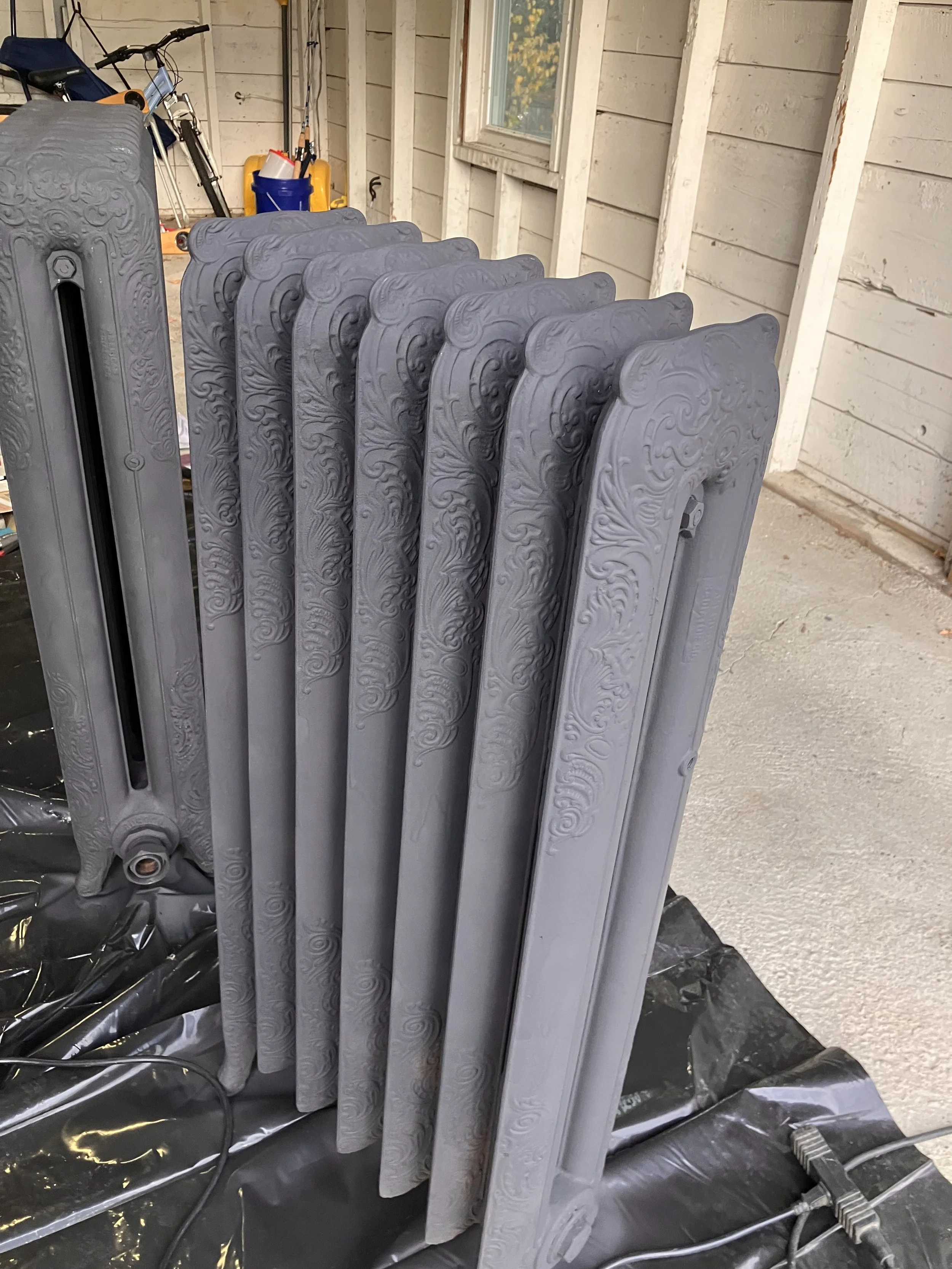 A row of freshly painted vintage cast iron radiators with intricate carvings, placed indoors on a black plastic sheet.