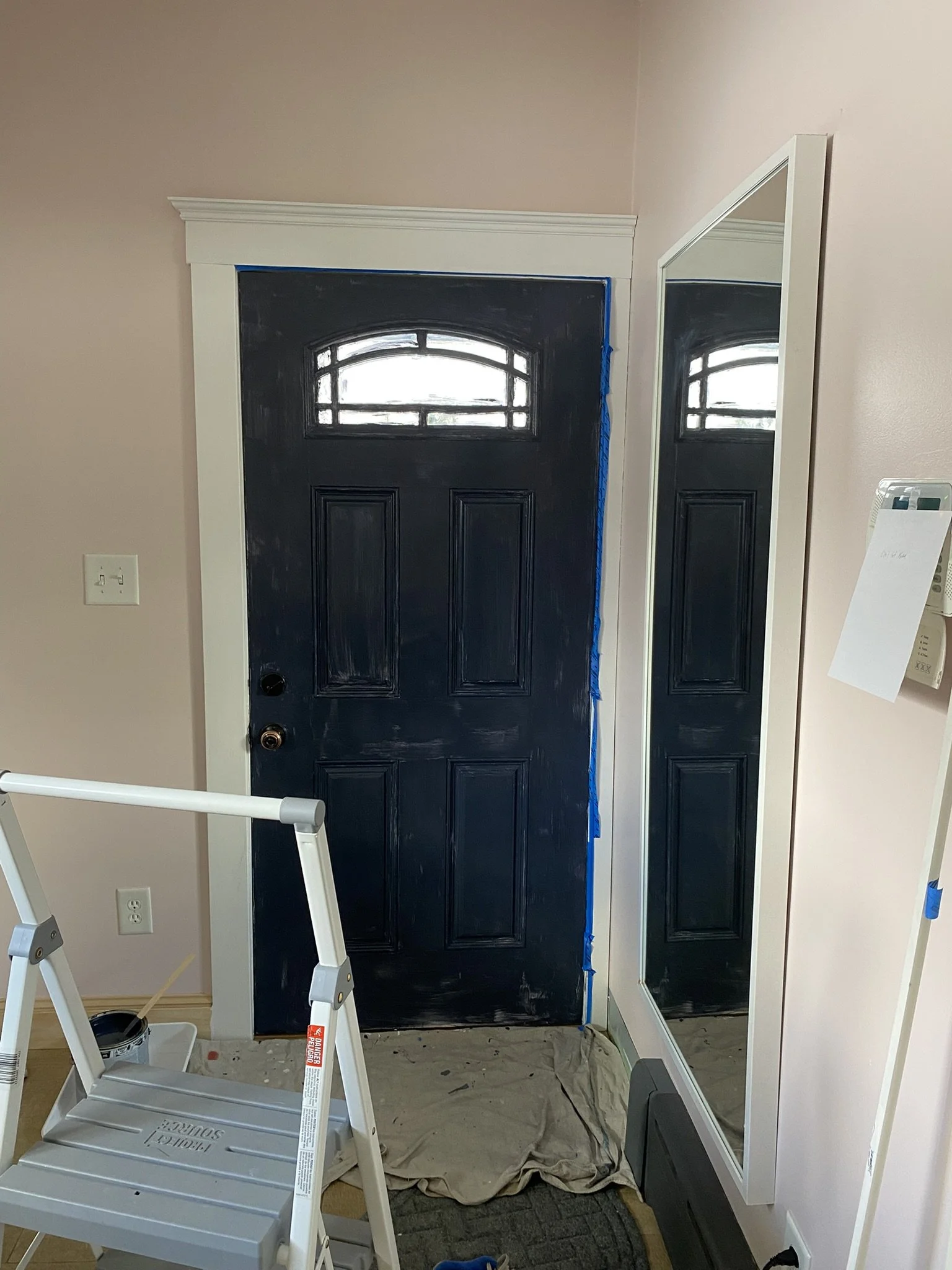 A black front door with a small arched window at the top, partially painted, with painter's tape on the sides. There is a large mirror leaning against the wall on the right, a step ladder in front, a paint can and cloth on the floor, and a beige wall