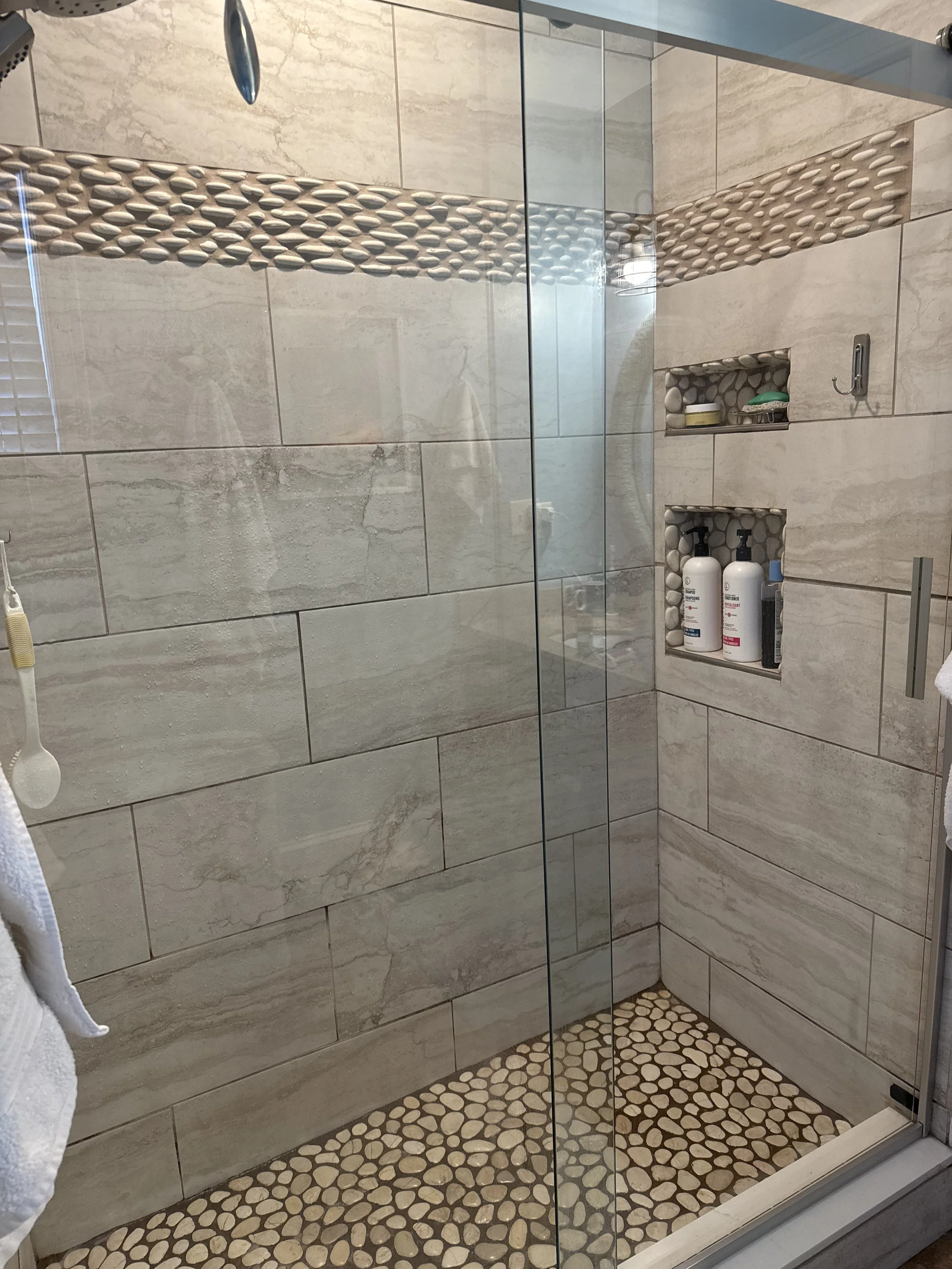 A tiled walk-in shower with glass enclosure, featuring pebble tile on the floor, a decorative pebble border, and built-in wall niches holding bottles and toiletries.