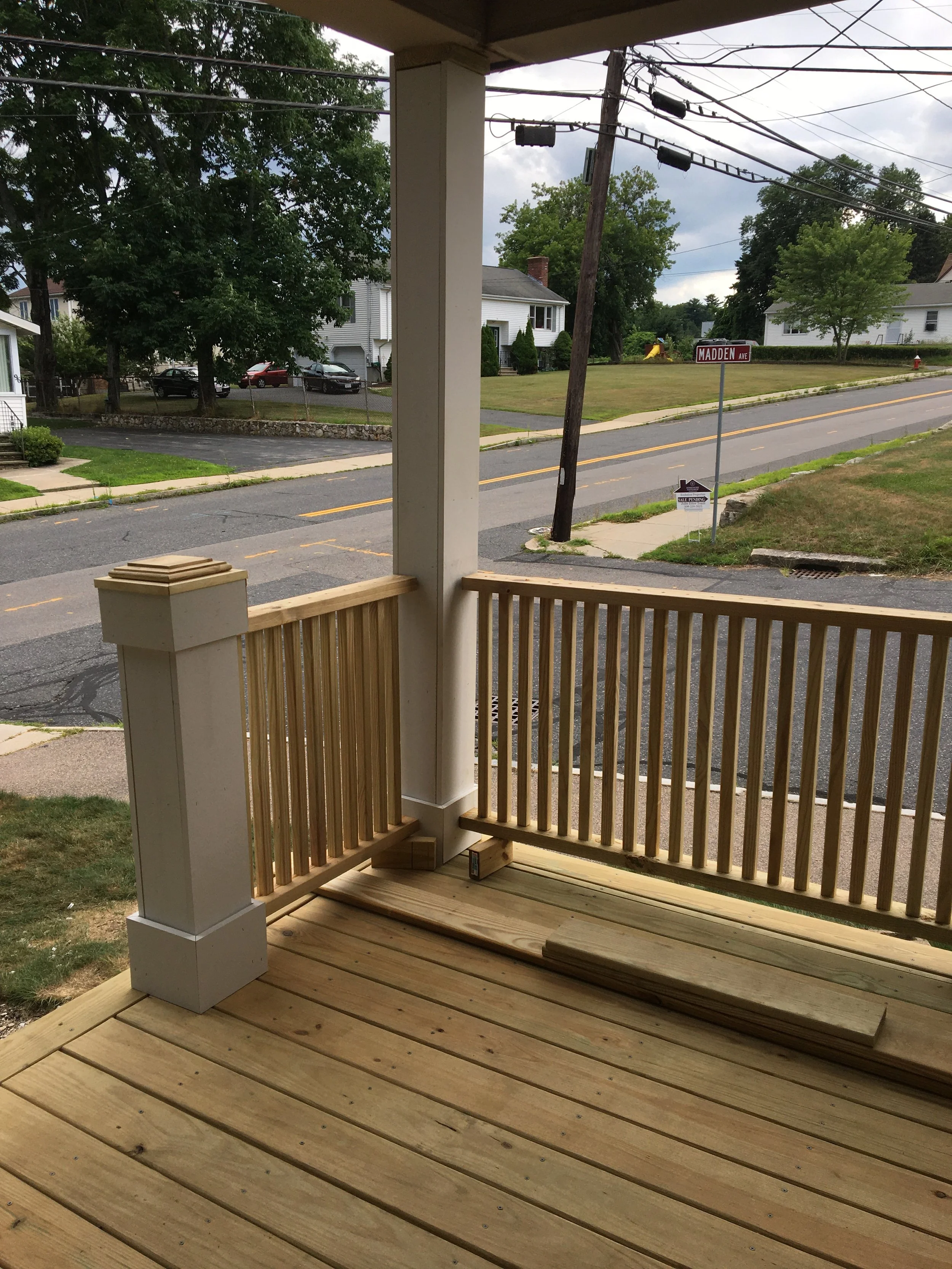 A front porch with new wooden decking, including a railing and some detached boards on the floor. The street and houses are visible in the background.