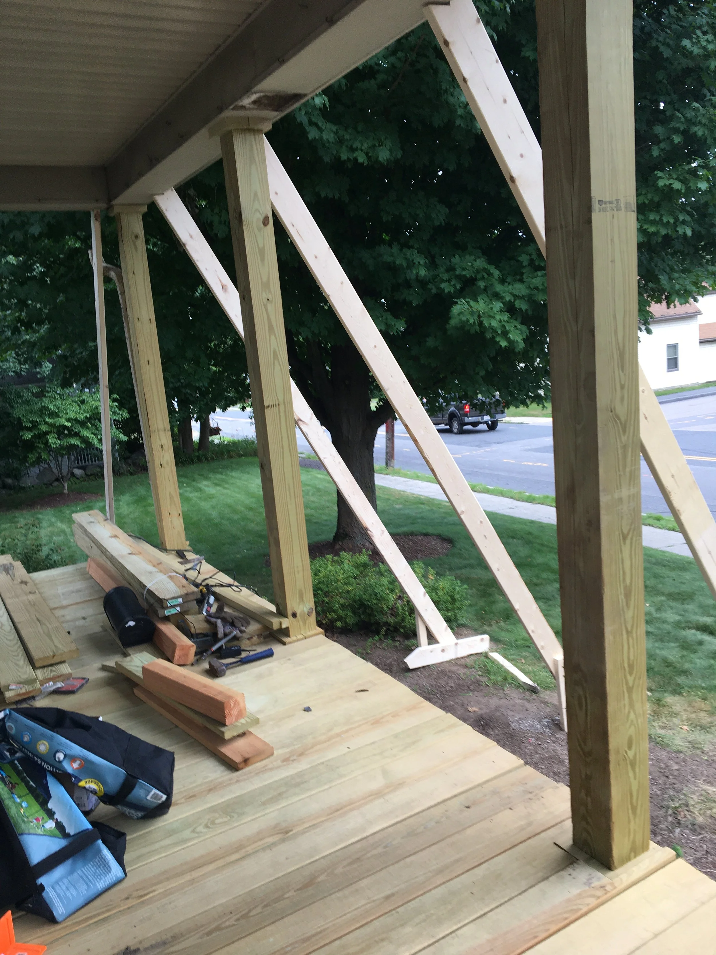 Wooden deck under construction with vertical and diagonal support beams, tools and materials on the floor, overlooking a green yard with a large tree and a street in the background.