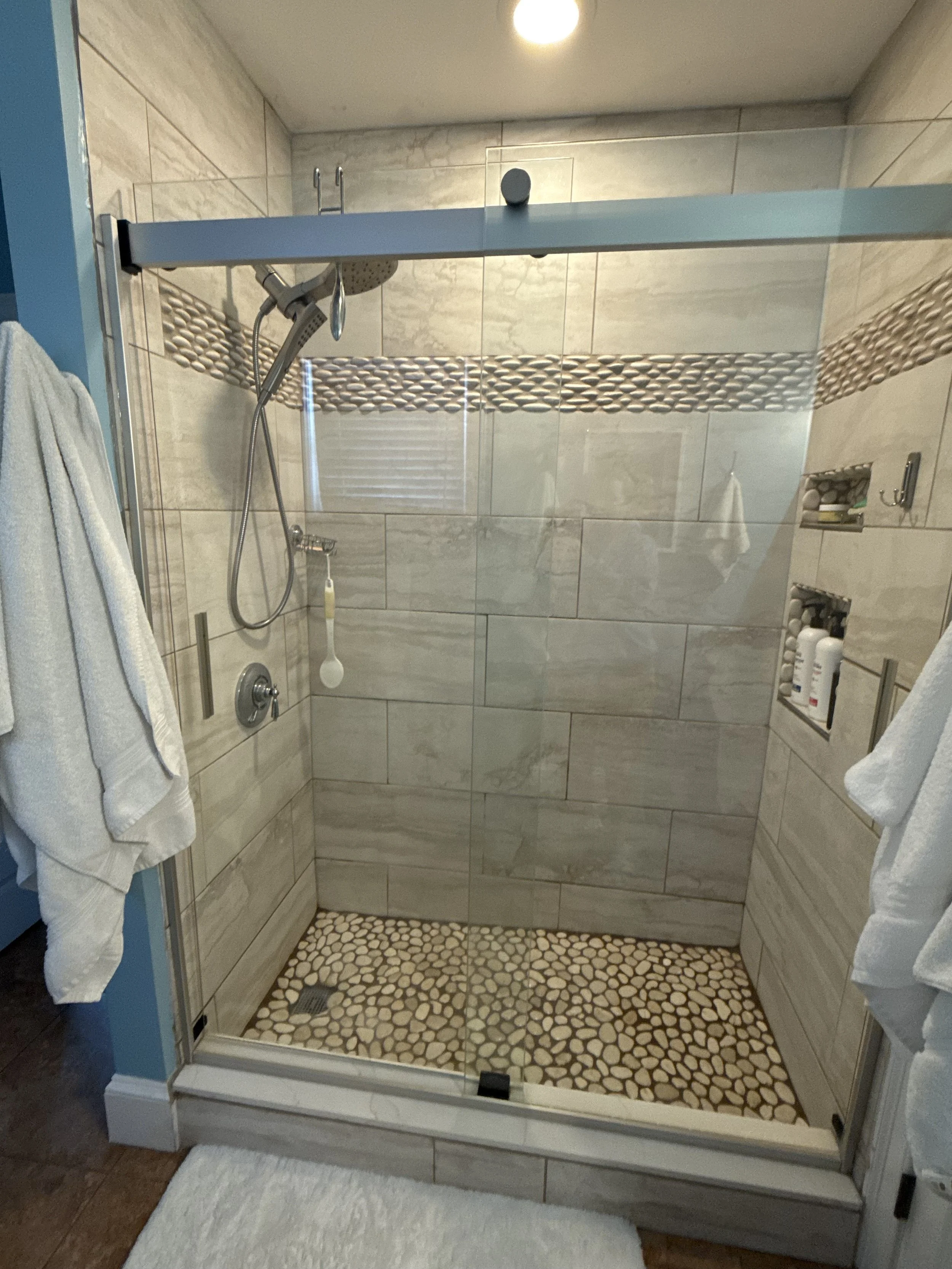 A modern walk-in shower with glass door, beige tiles, pebble flooring, built-in niche with toiletries, showerhead, and handheld sprayer.