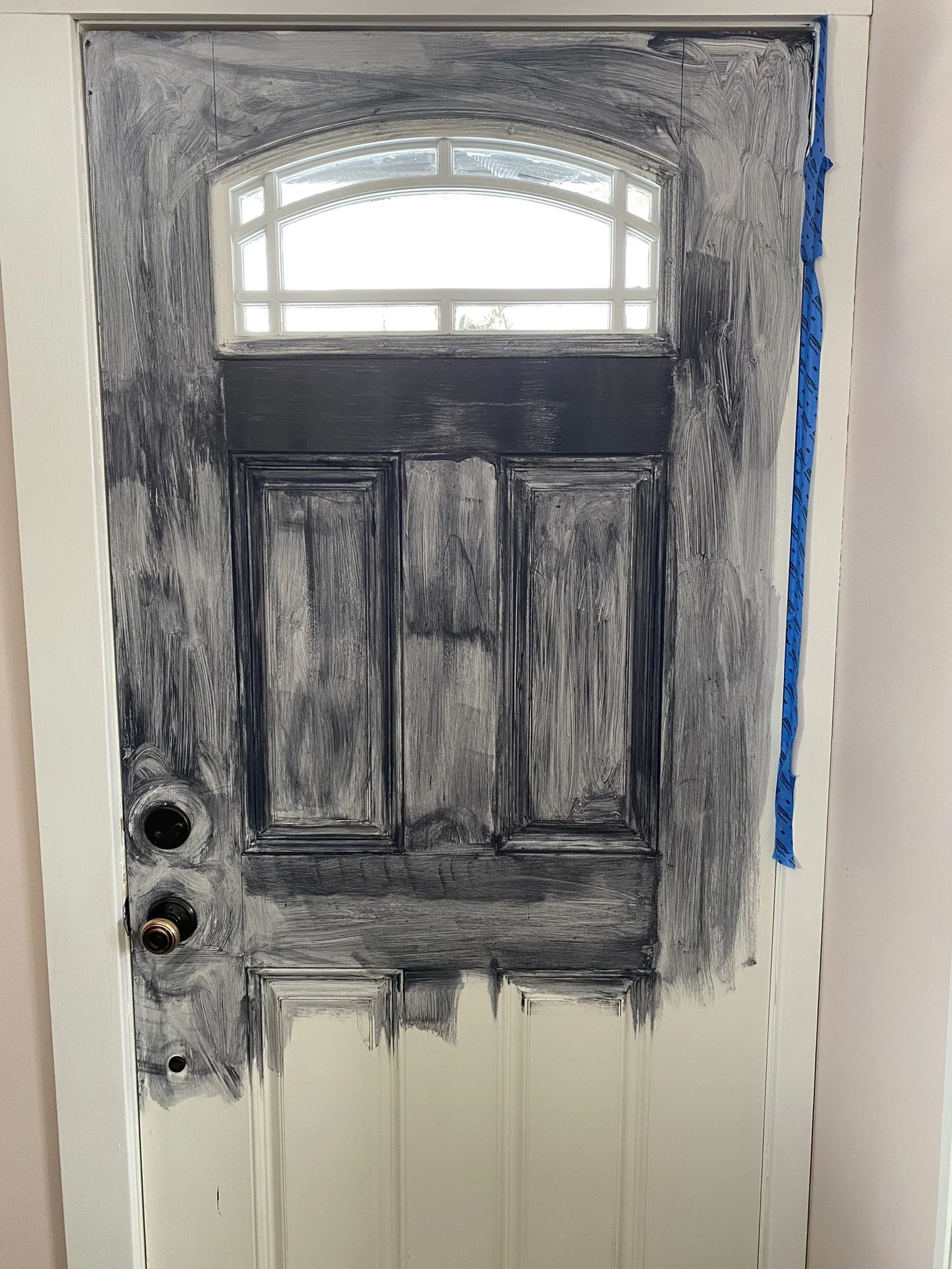 A front door with black paint being sanded down, revealing the white surface underneath, with a window at the top and blue painter's tape on the right side.