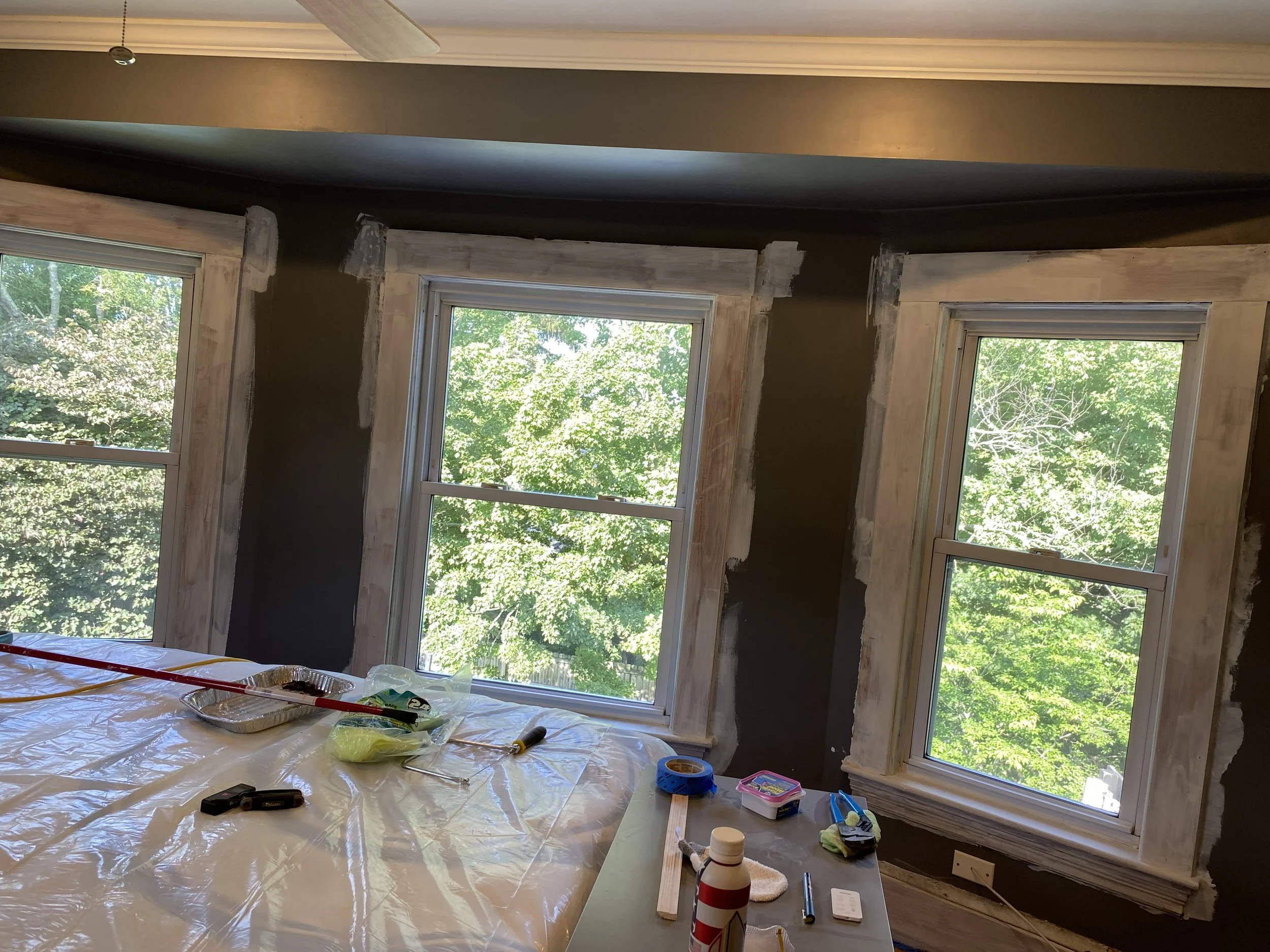Room under renovation with three windows, painter's tape, tools, and supplies on a table.