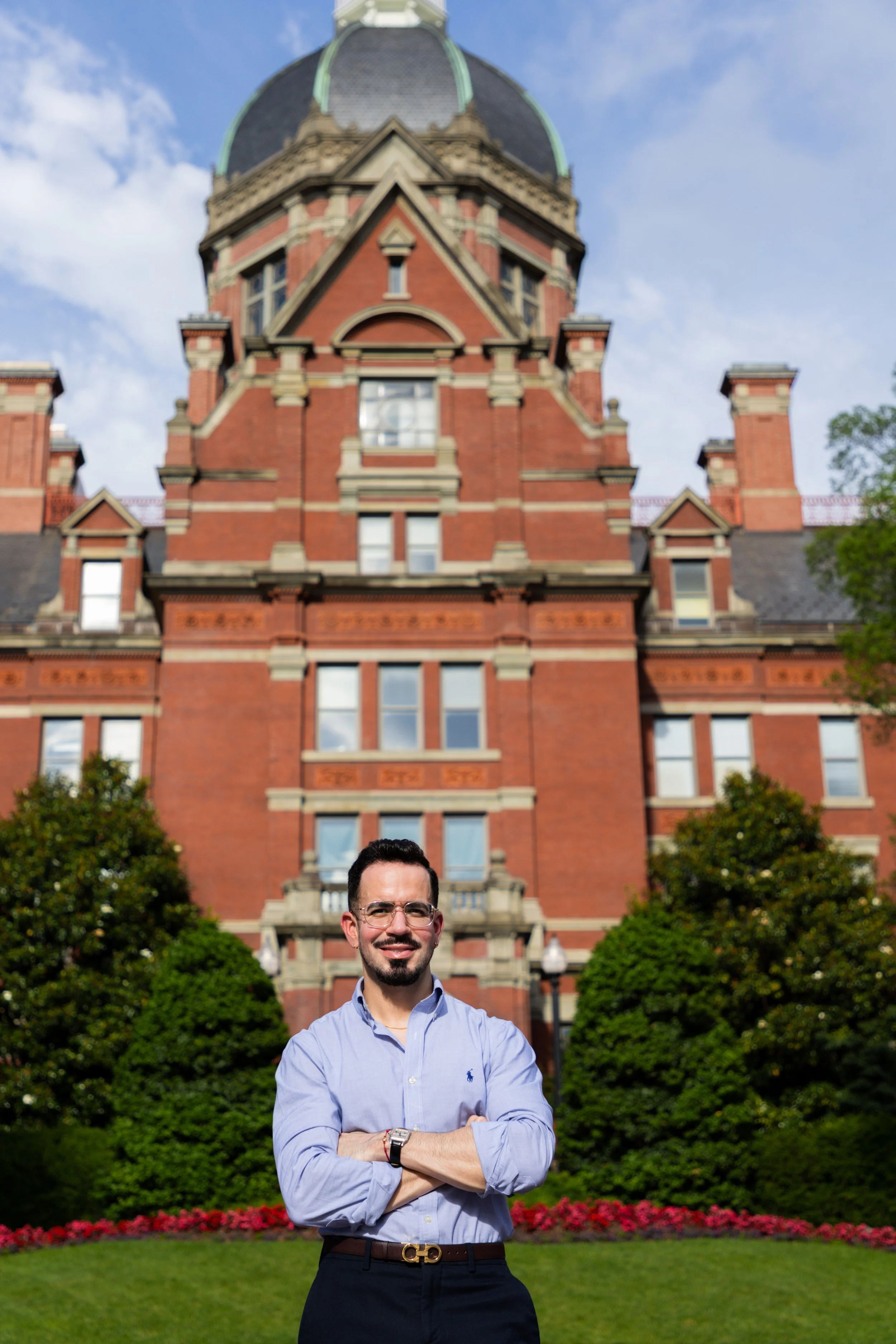 A man with glasses and a beard wearing a light blue button-down shirt and dark pants, standing outdoors with his arms crossed, in front of a red brick historic building with a large dome and towers, some trees, and a neatly maintained lawn with red f