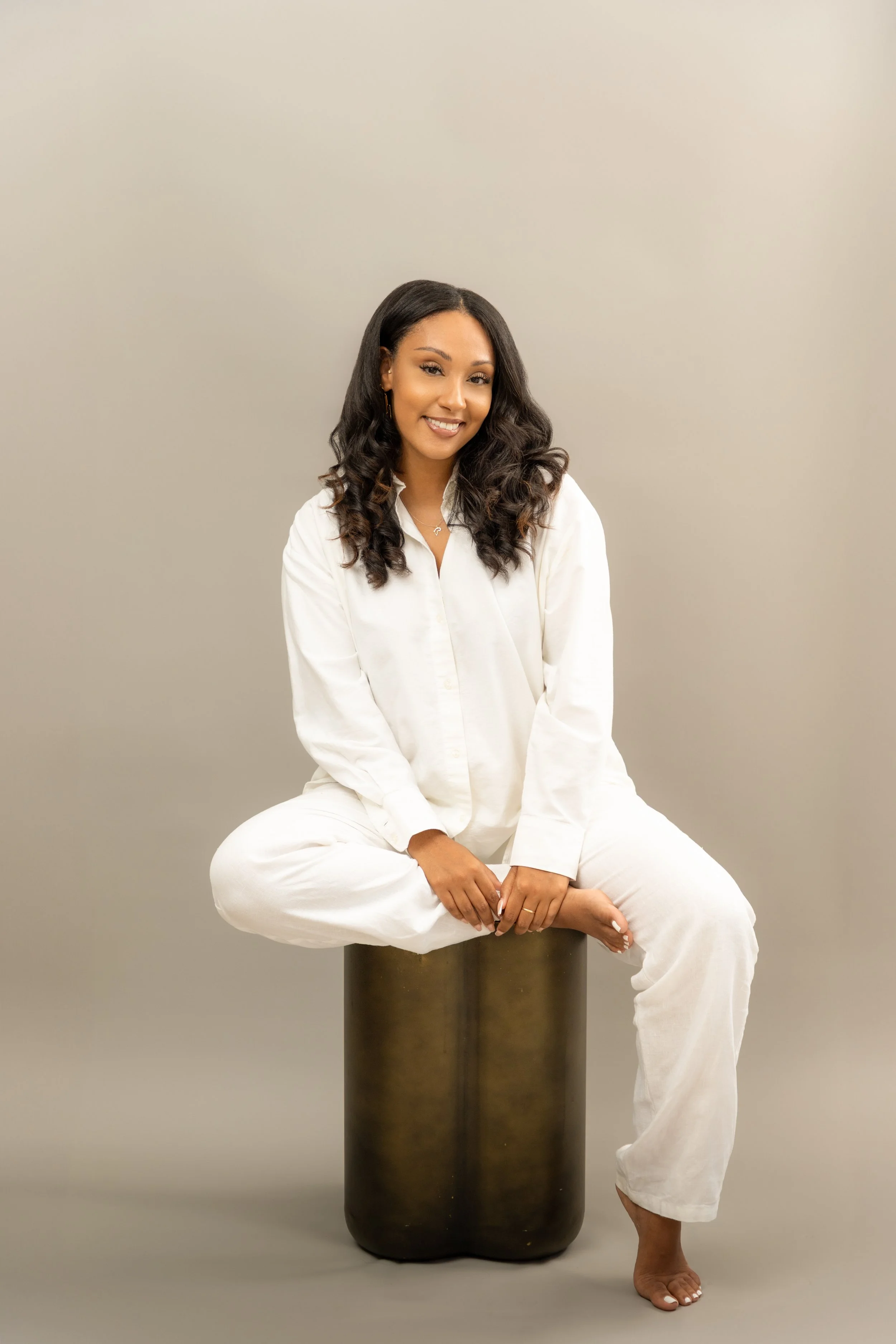 A young woman with dark wavy hair, dressed in a white shirt and white pants, sitting cross-legged on a cylindrical black stool against a plain beige background, smiling at the camera.