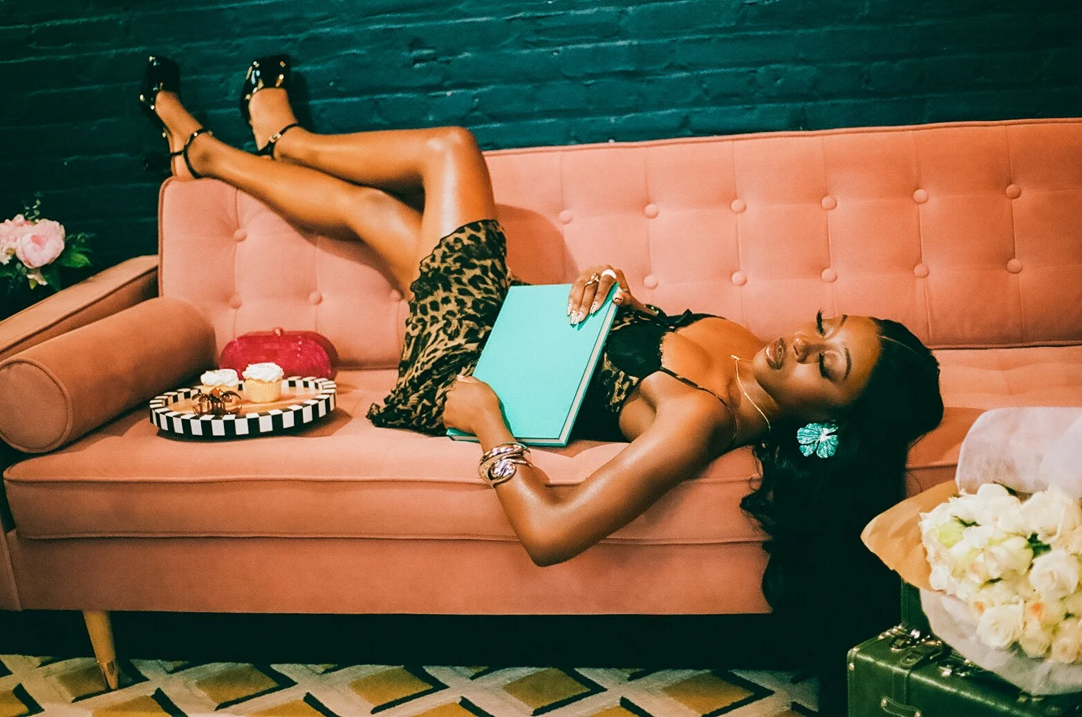 A woman lying on a pink sofa, wearing a leopard print dress and black high heels, holding a closed teal book, with a tray of cupcakes and a bouquet of white roses nearby.