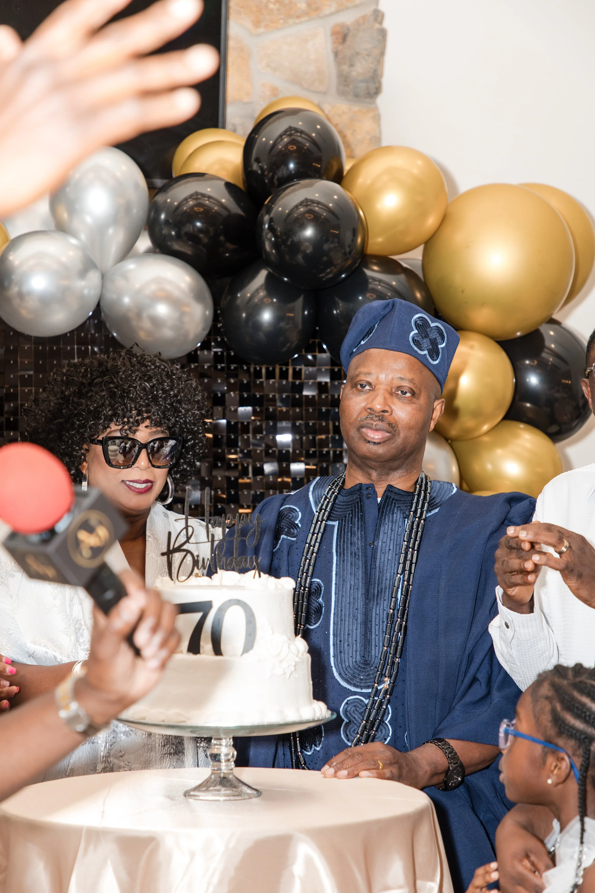 Guest of honor in traditional attire cutting a birthday cake in front of a gold balloon arch