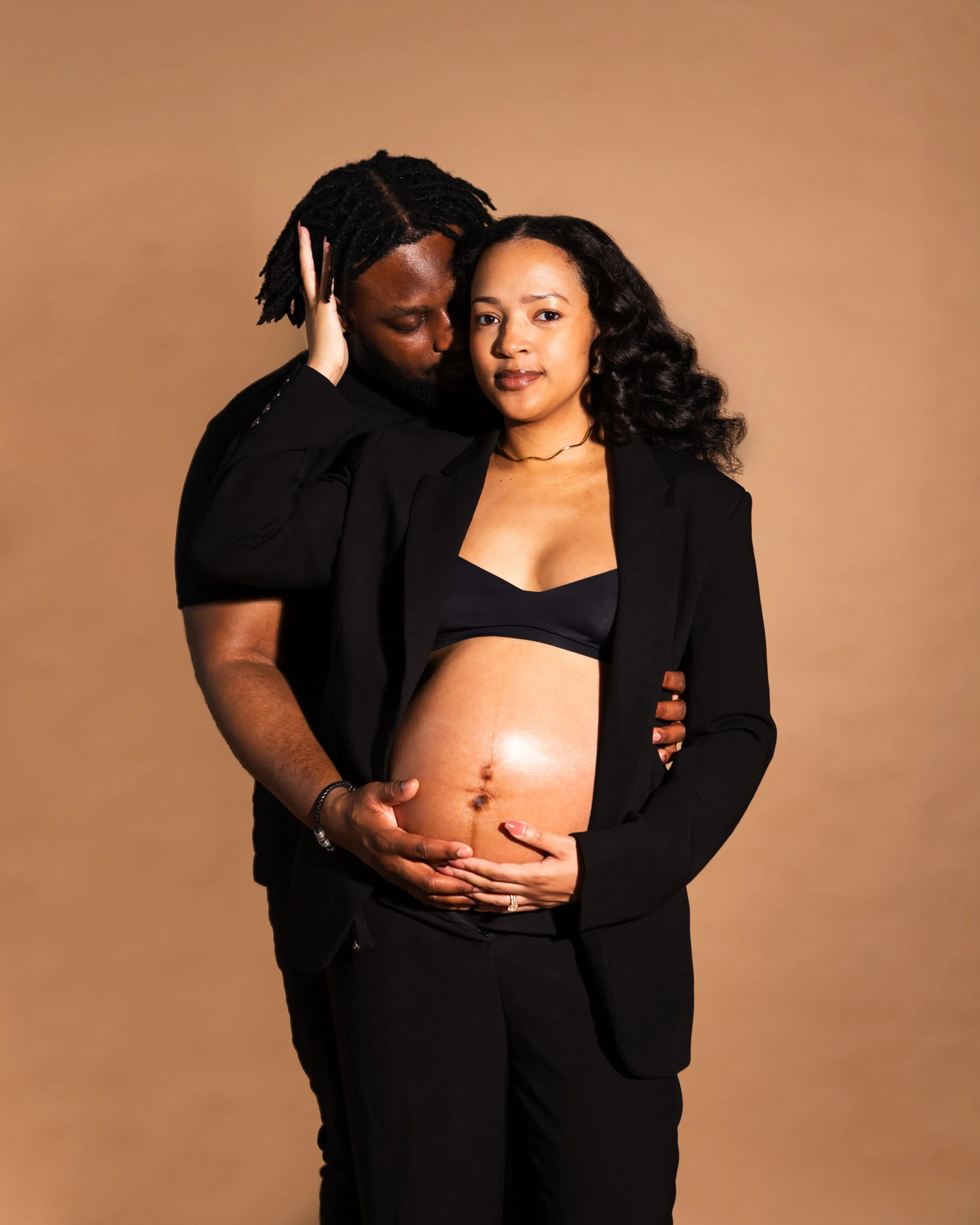 A pregnant woman in a black blazer and top, with curly hair, is holding her belly, while a man with dreadlocks, dressed in black, stands behind her with his hand on her stomach and his face close to her head, on a plain beige background.