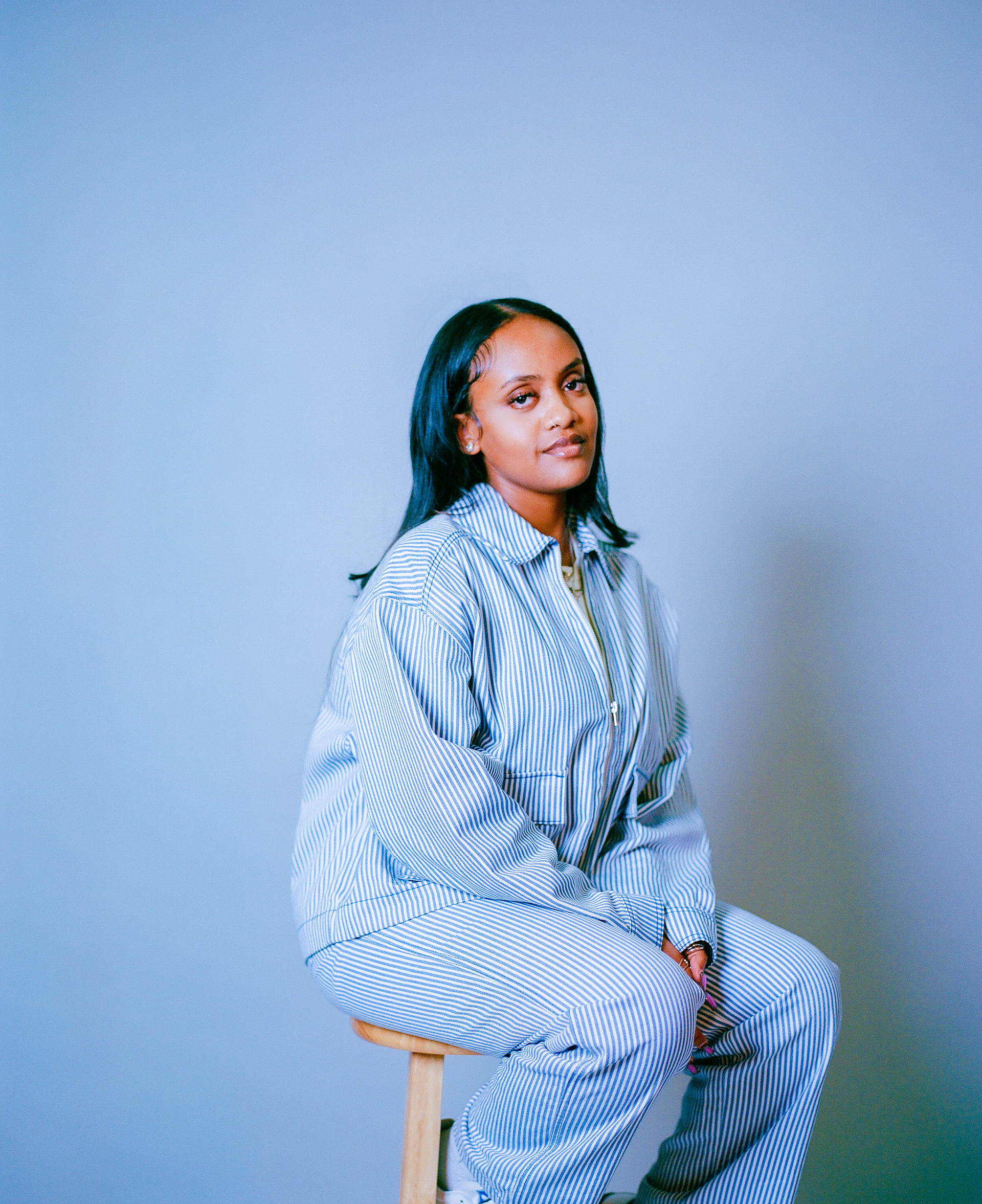 A woman with dark hair wearing a blue and white striped jacket and matching pants, sitting on a wooden stool against a plain light-colored background.