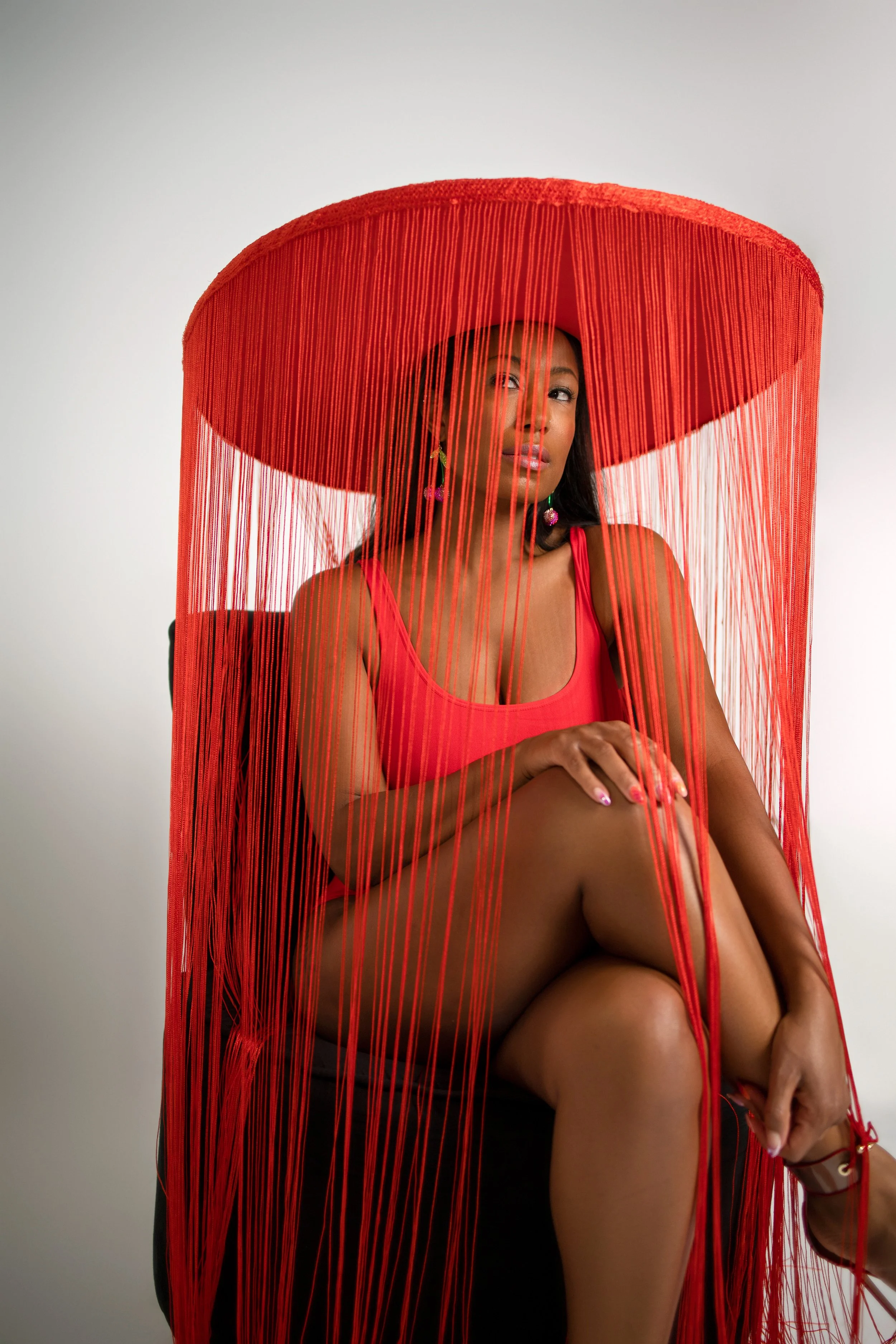 Woman wearing a large red hat with hanging fringe, red dress, sitting with legs crossed against a white background.