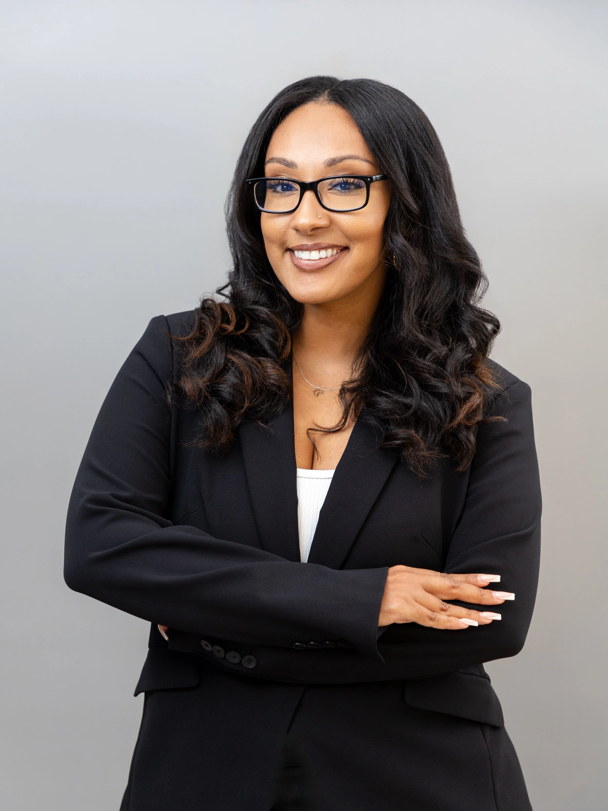 A woman with black, curly hair, glasses, and a black blazer smiling with arms crossed against a neutral background.
