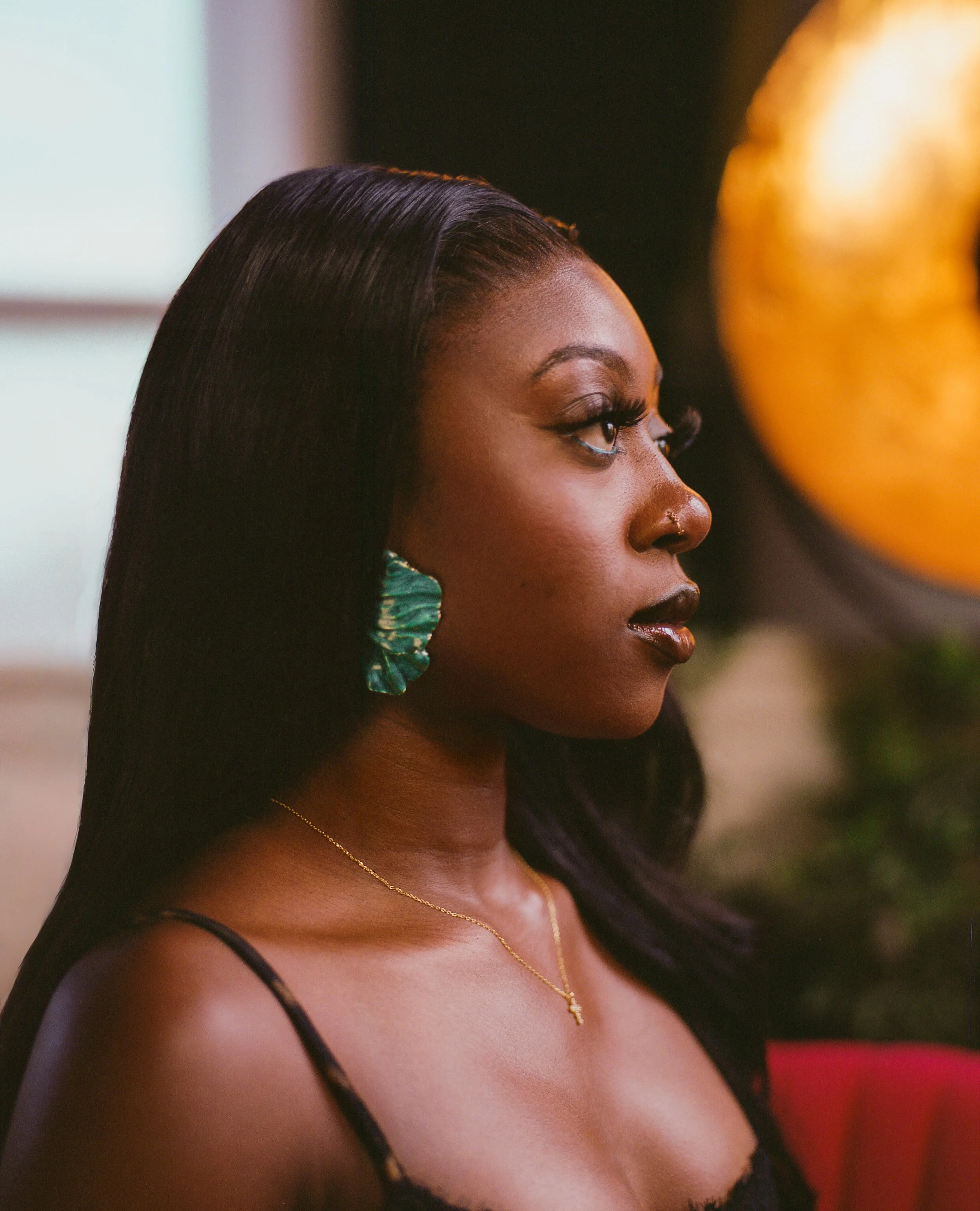 A woman with long black hair, wearing green earrings, a gold necklace, and a black top with straps, sitting in profile with a blurred background.