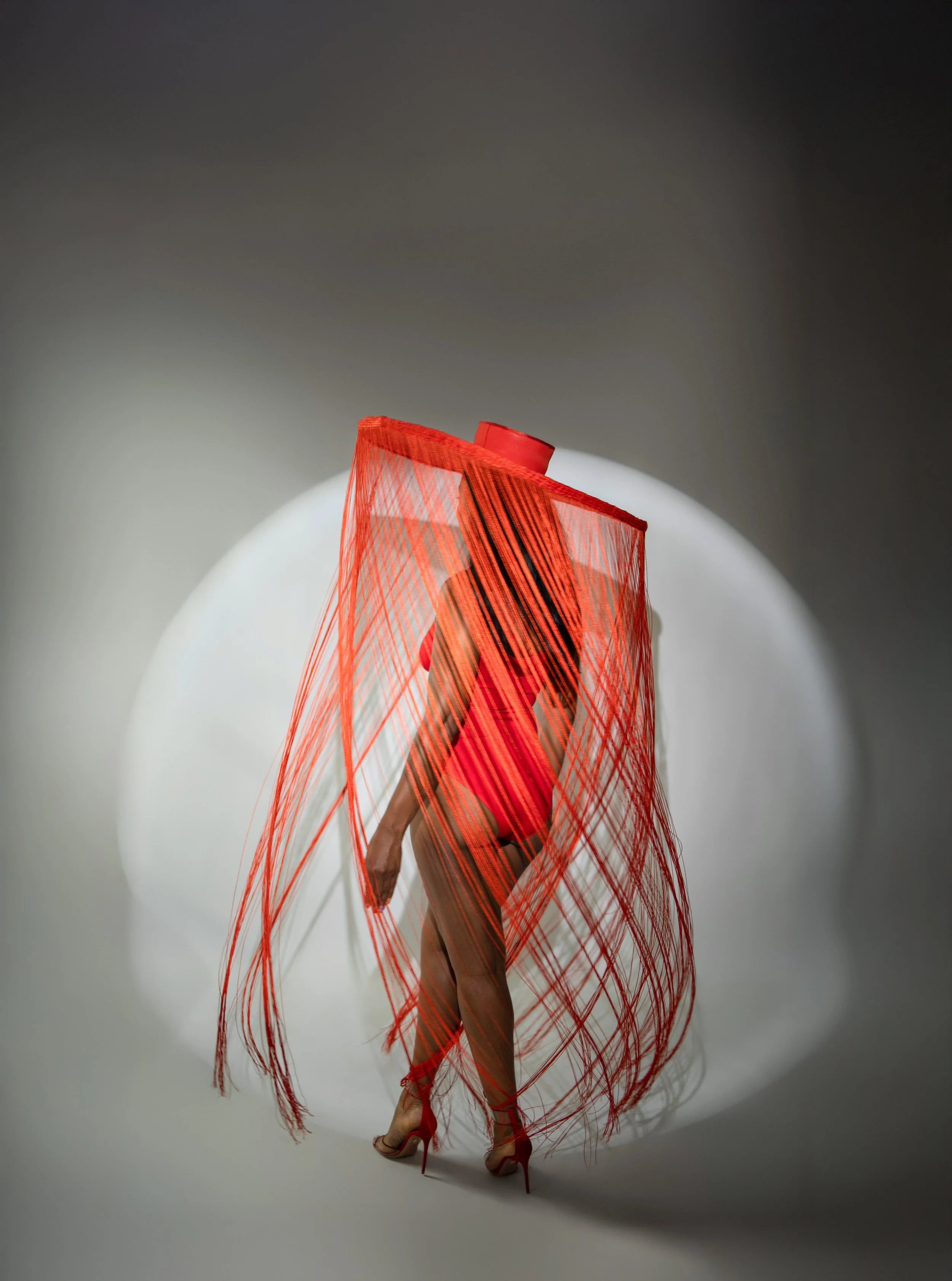 A model wearing a red dress with sheer fabric and high heels, standing behind a red mesh curtain, against a plain background with a spotlight effect.