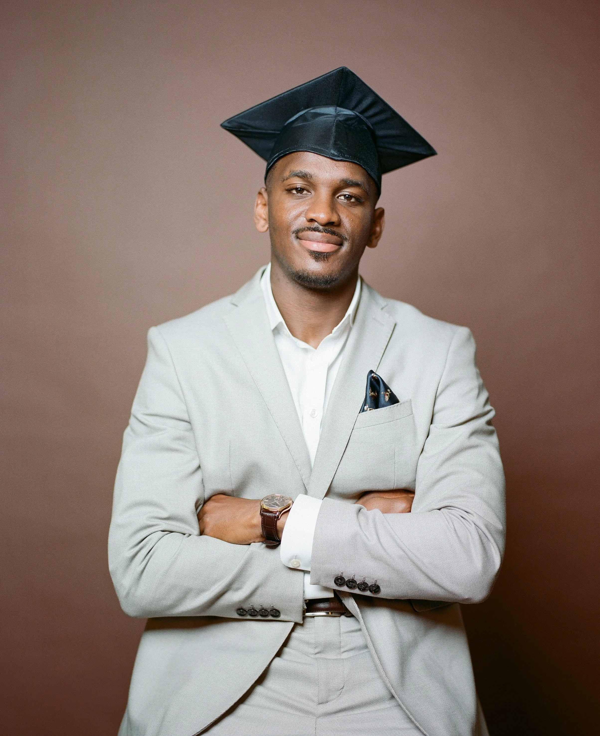 A young man in a light gray suit with a white shirt and pocket square, wearing a black graduation cap, standing with arms crossed against a brown background.