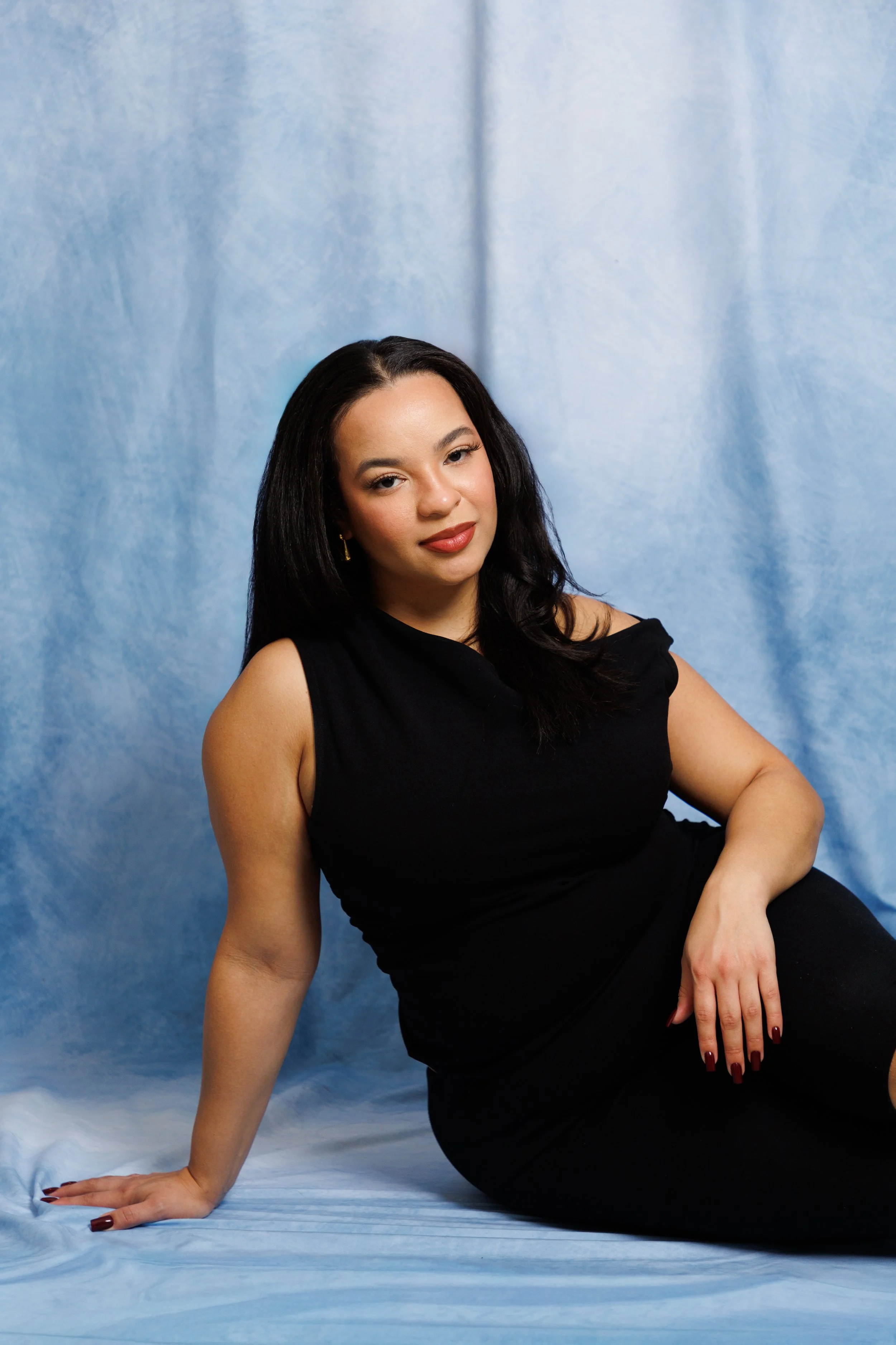 A woman with long black hair, wearing a sleeveless black dress, sitting on a blue fabric backdrop.