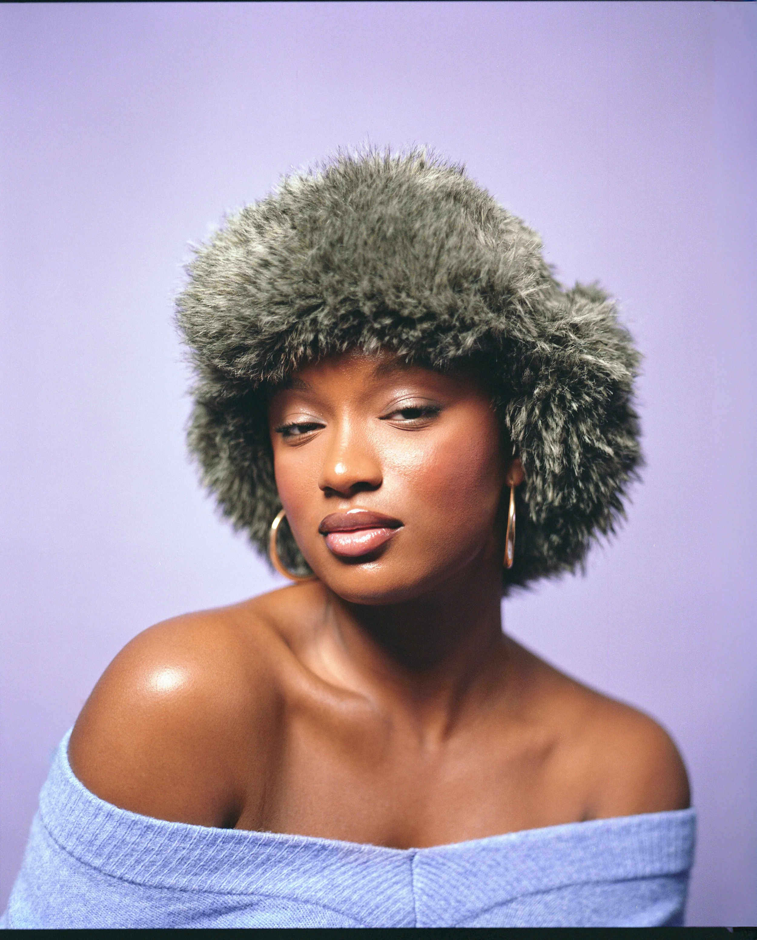 A portrait of a woman with dark skin, wearing a large fur hat, hoop earrings, and a light purple off-the-shoulder top, against a purple background.