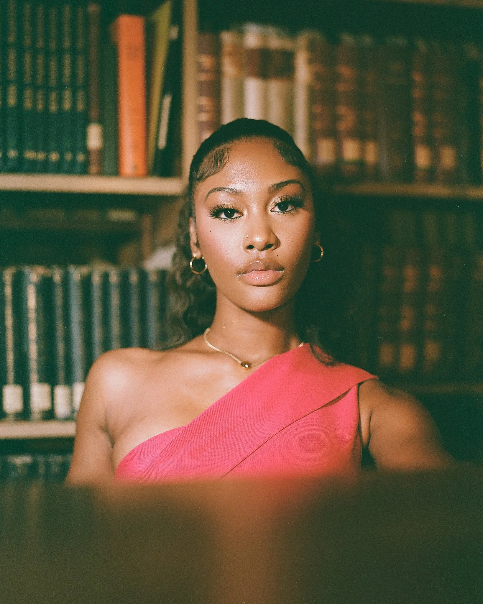 A young woman with dark curly hair pulled back, wearing gold hoop earrings, a necklace, and a pink off-shoulder top, standing in front of a bookshelf filled with books.