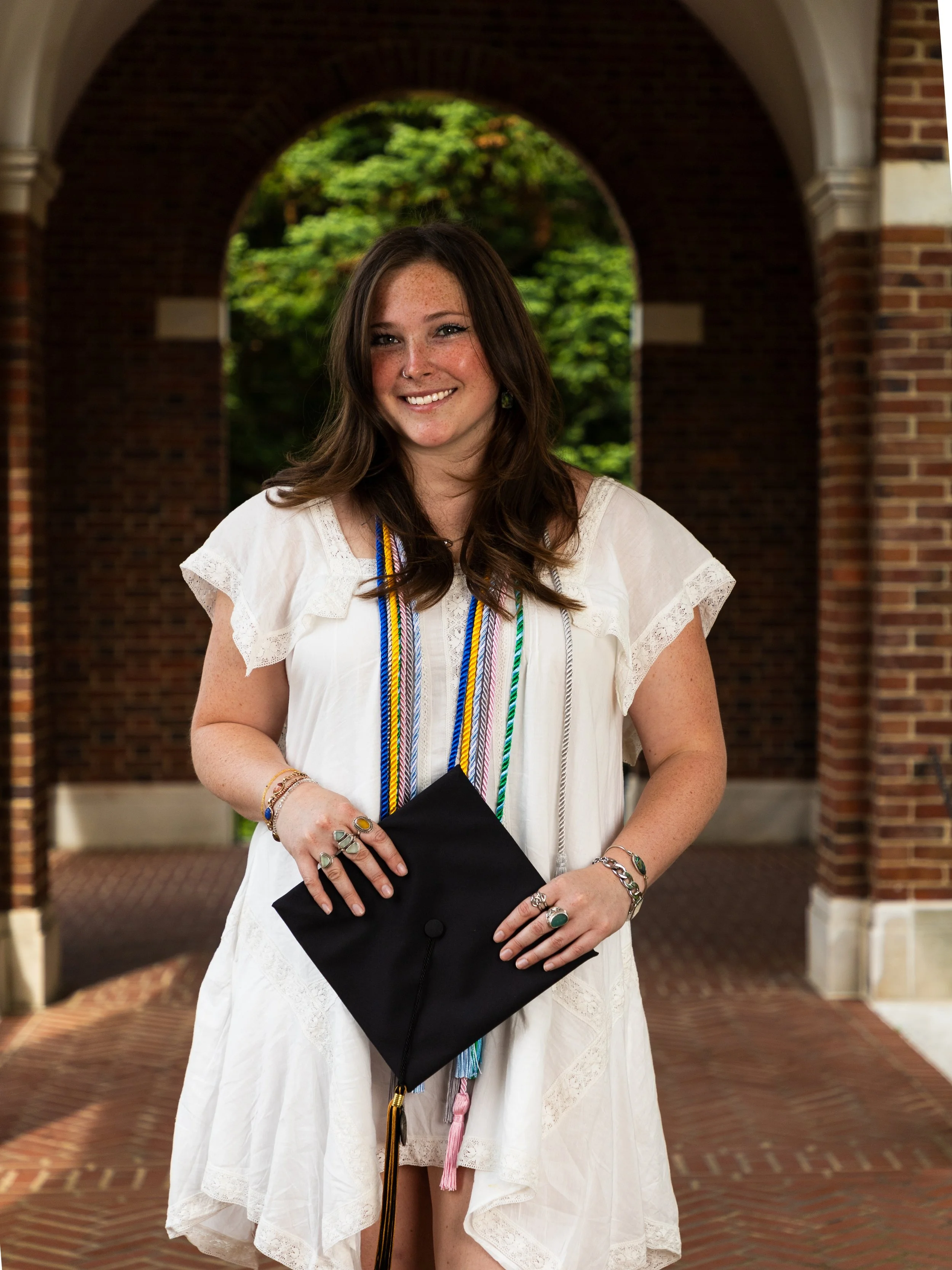 A young woman in a white dress holding a black graduation cap, standing under an archway with brick walls and greenery in the background.