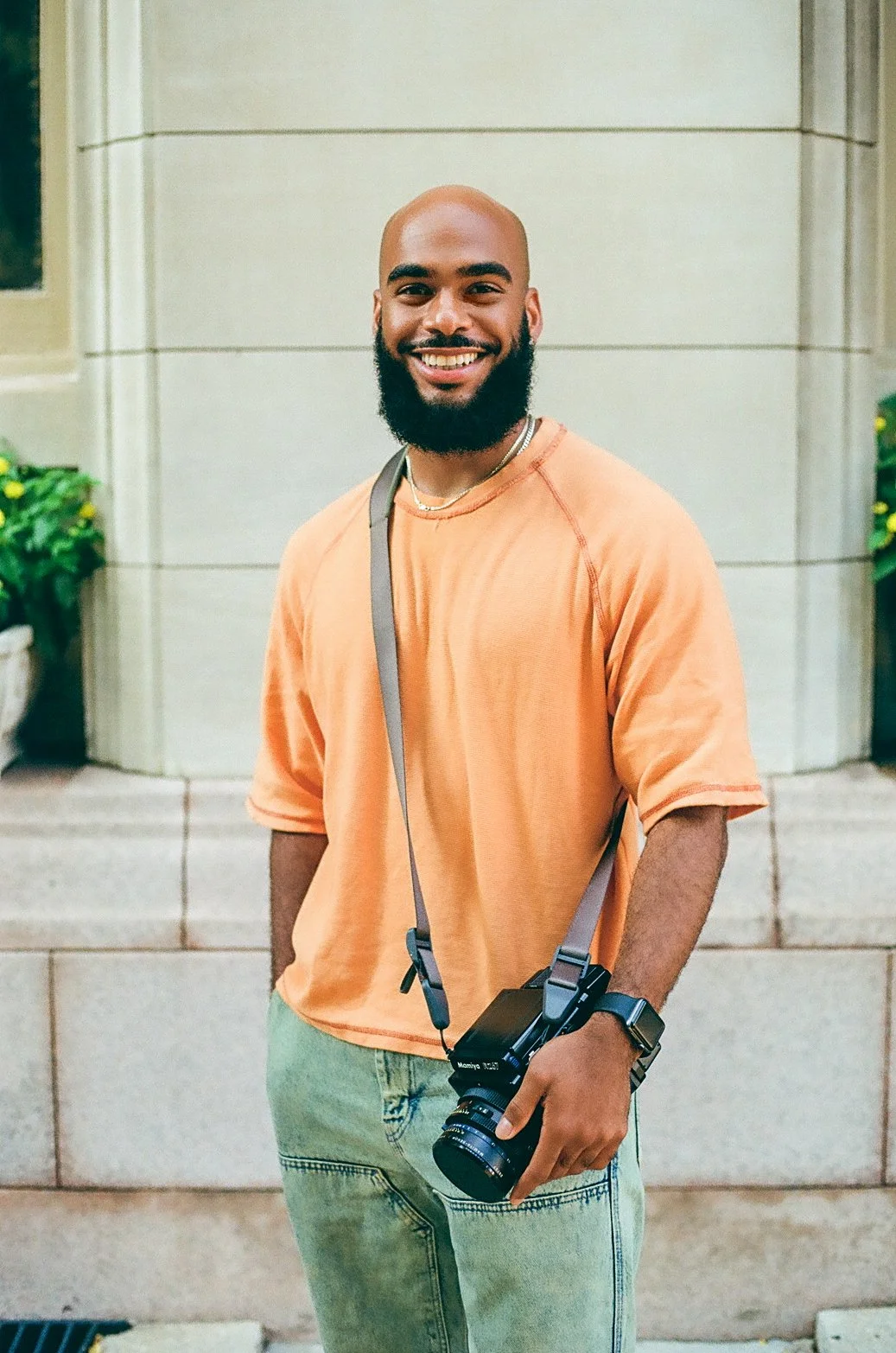 Smiling man with a beard, wearing an orange T-shirt, jeans, and a watch, holding a camera with a strap around his neck, standing outdoors in front of a building with potted plants nearby.