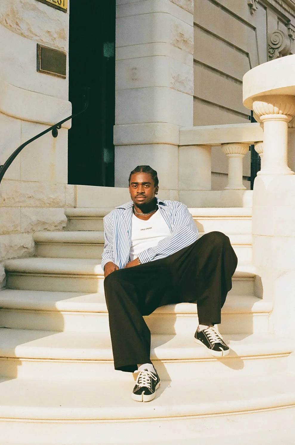 A man sitting on white marble steps outside a building with classical architectural details, wearing a striped shirt, white t-shirt, black wide-leg pants, and sneakers.