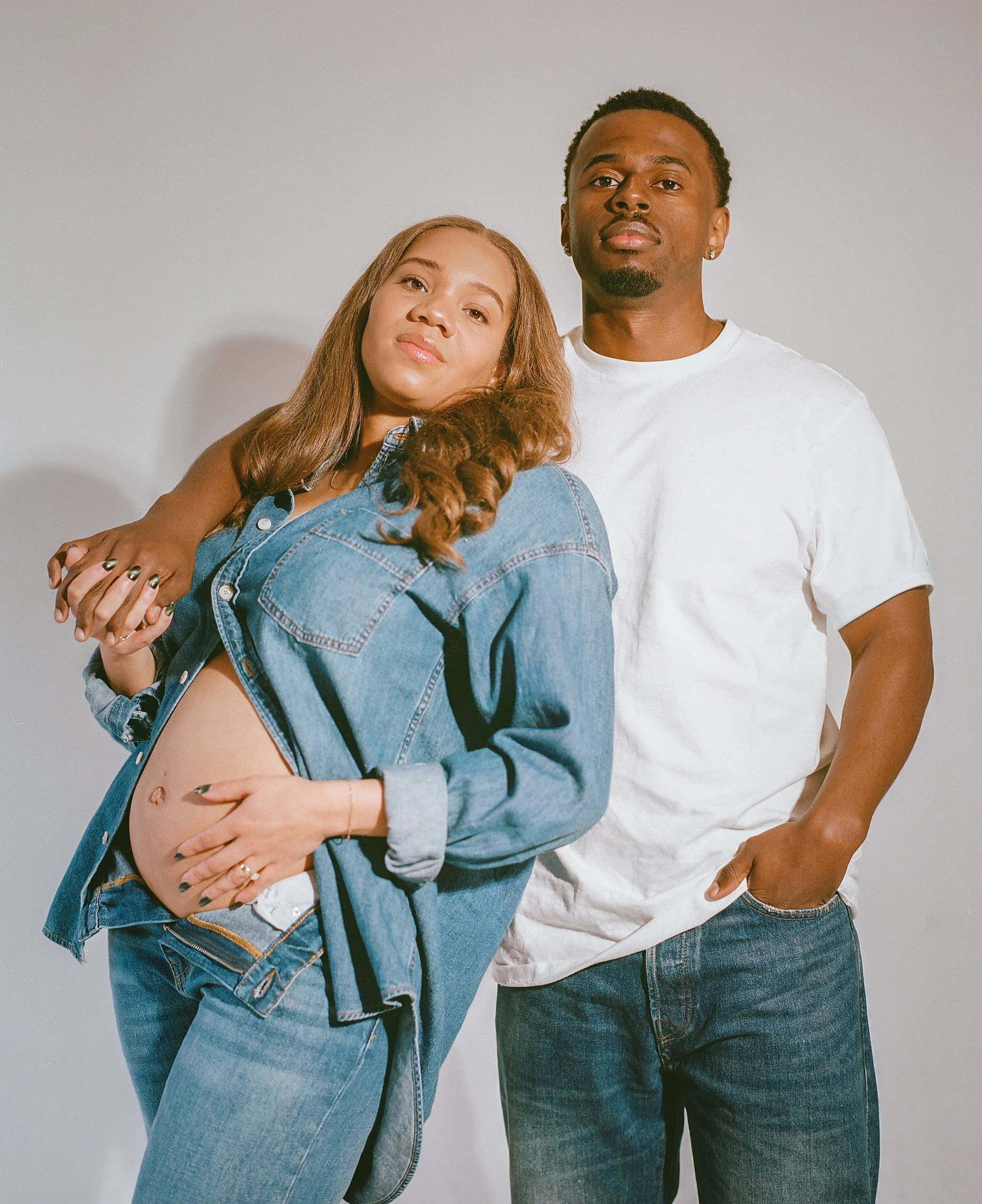 A pregnant woman and a man standing together against a plain background. The woman is wearing a denim jacket and jeans, with her hand on her belly, and the man is wearing a white t-shirt and jeans, with one hand in his pocket.