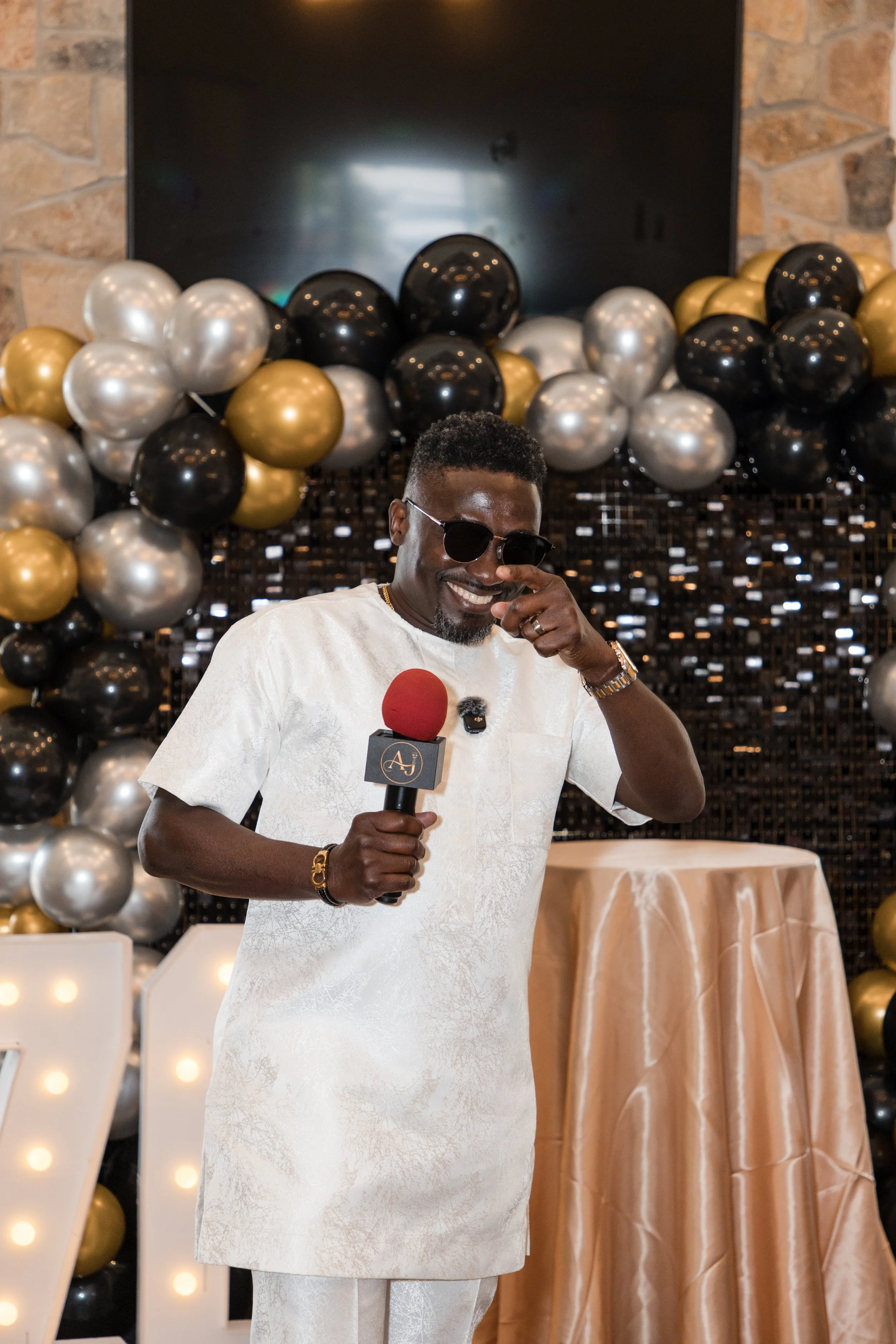 Host in white speaking into a microphone before a black and gold balloon backdrop at a birthday celebration