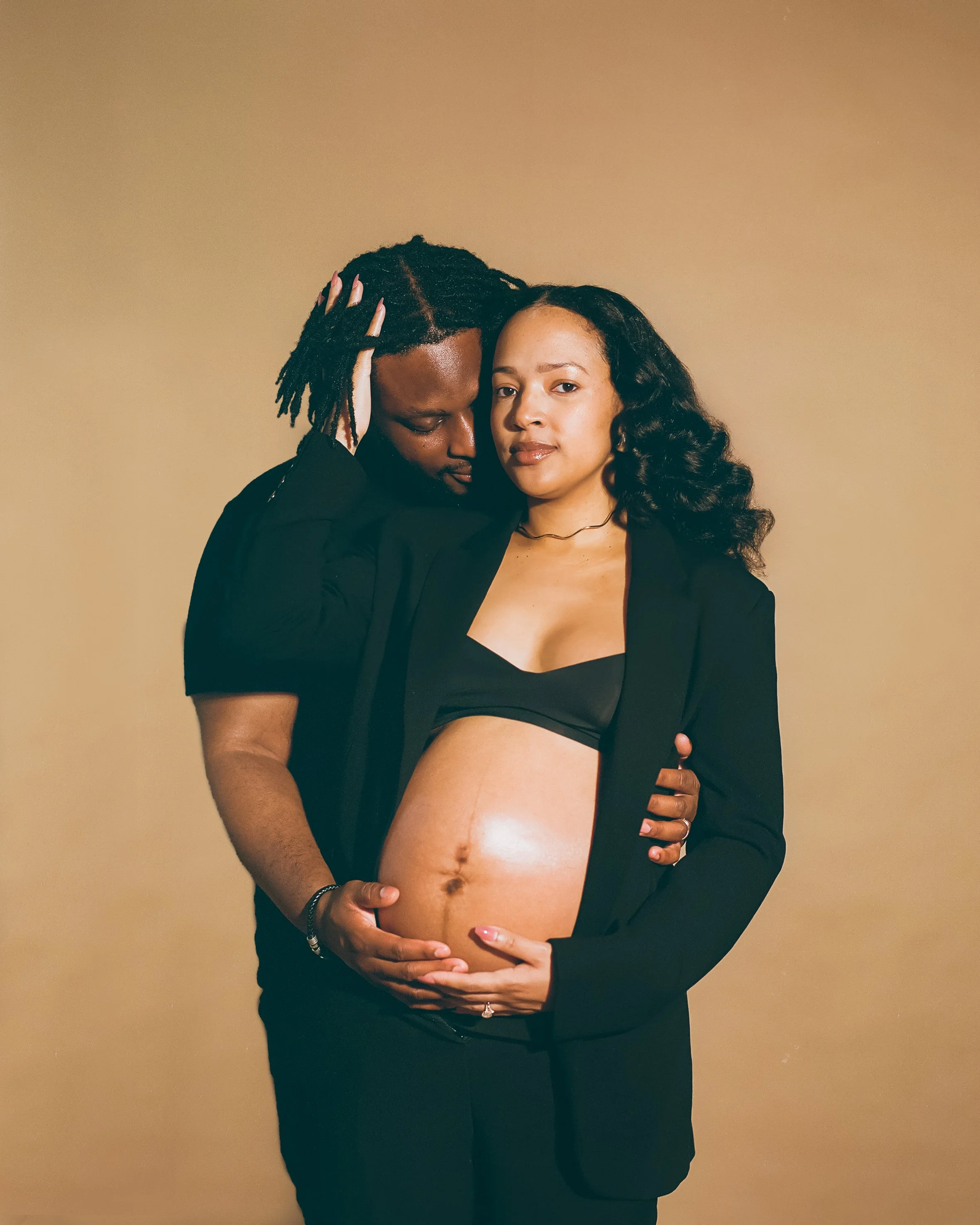 A pregnant woman with curly hair and a black top showing her belly, wearing a black blazer, standing in front of a beige background. A man with dreadlocks and a black shirt, standing behind her, cradles her belly and holds her face gently, both looki