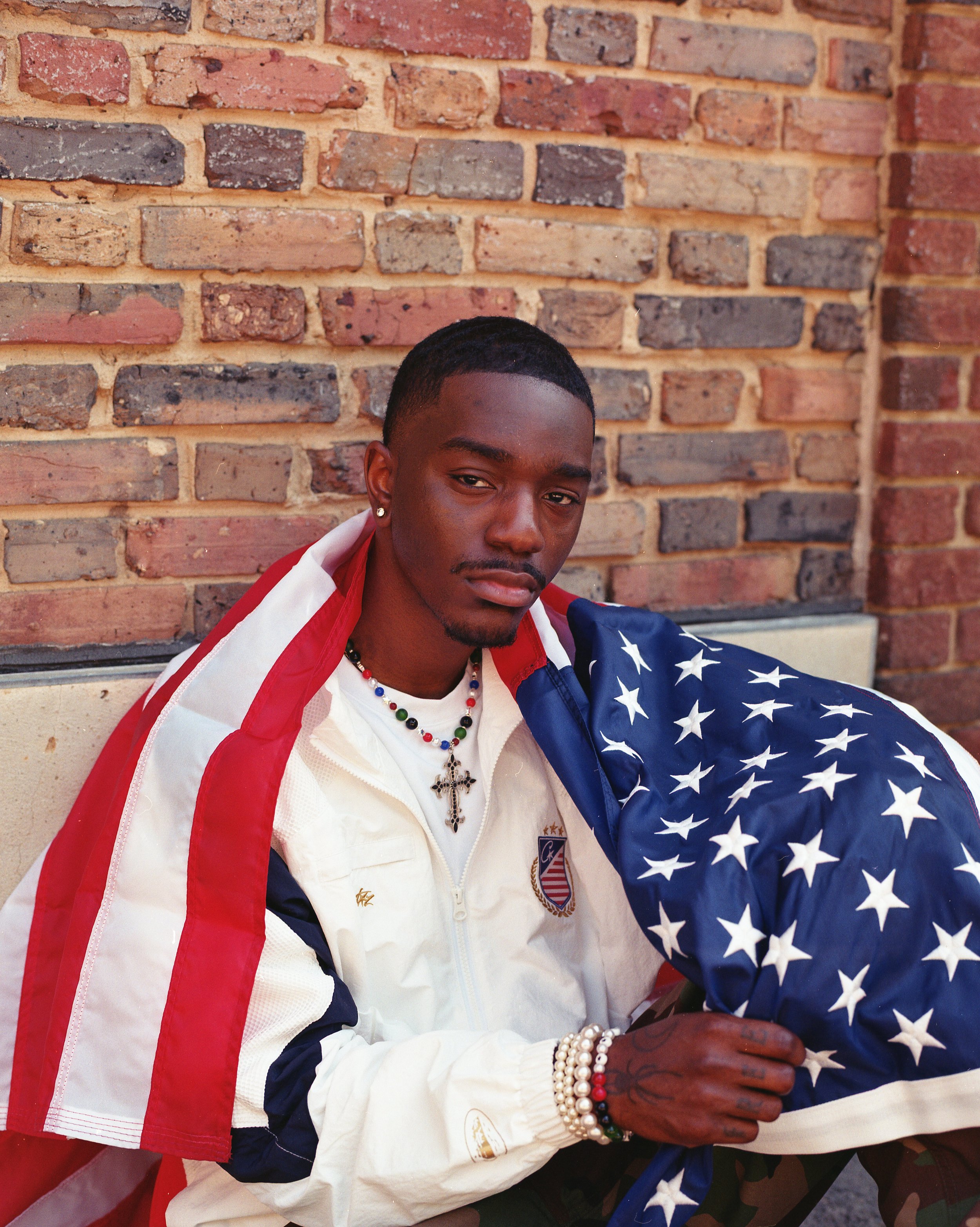 A young man with dark skin, wearing a white jacket with red and blue accents, adorned with jewelry including a cross necklace and pearl bracelets, sits against a brick wall. He holds a folded American flag with stars visible.