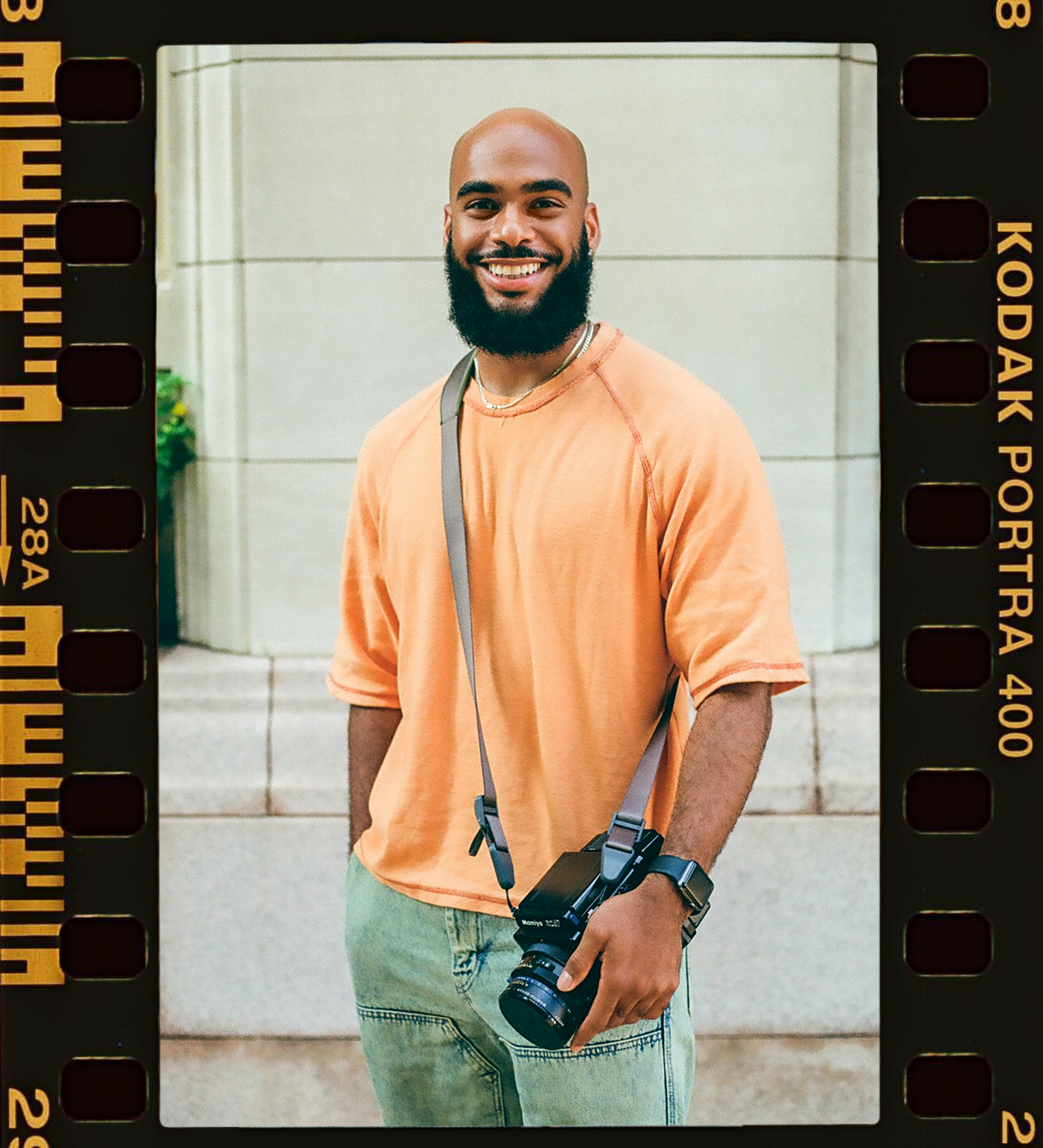 Smiling man with a camera hanging from his neck, wearing an orange T-shirt and light jeans, standing in front of a light-colored building.
