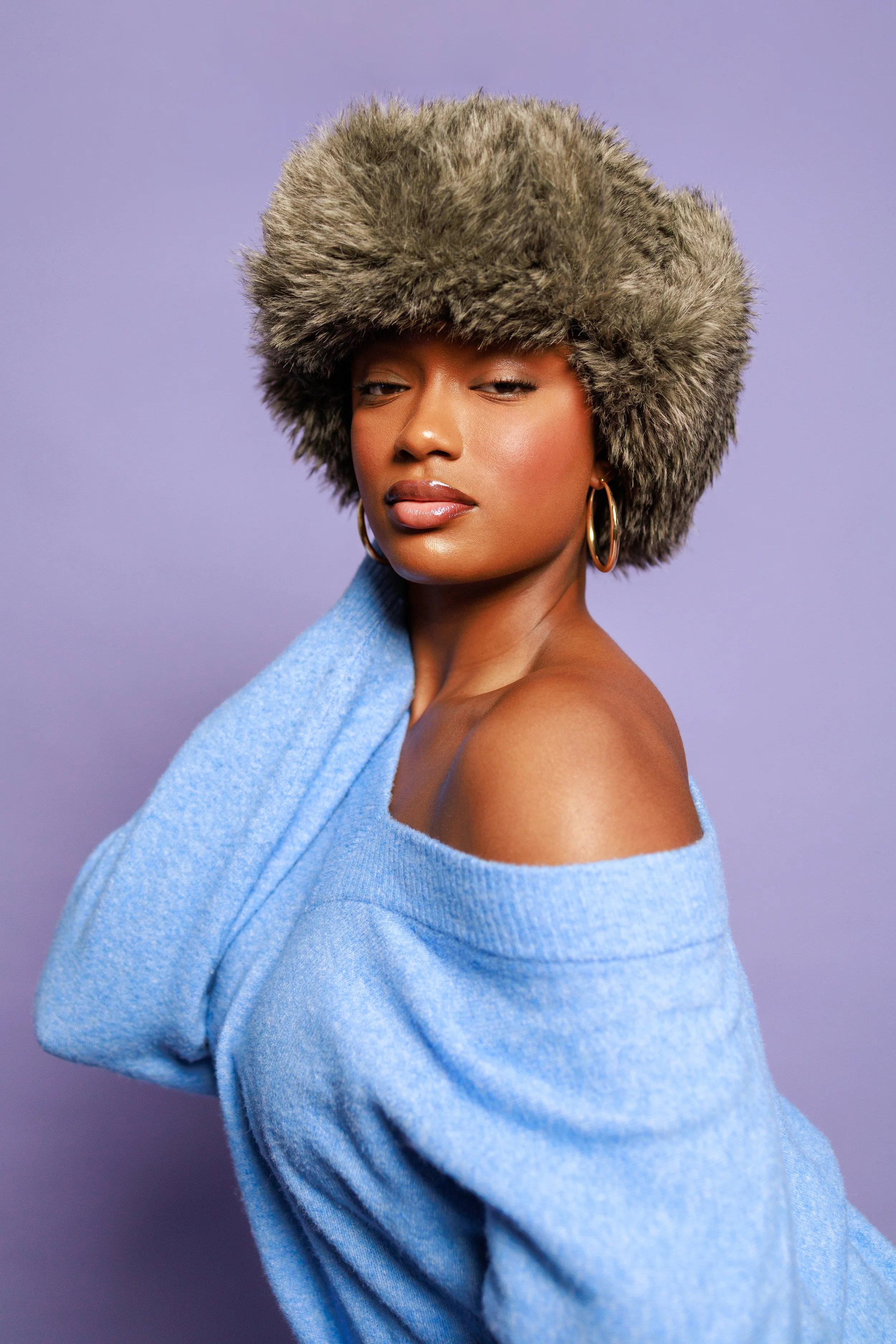 A woman with medium brown skin wearing a large fur hat, gold hoop earrings, and a cozy off-the-shoulder blue sweater against a purple background.