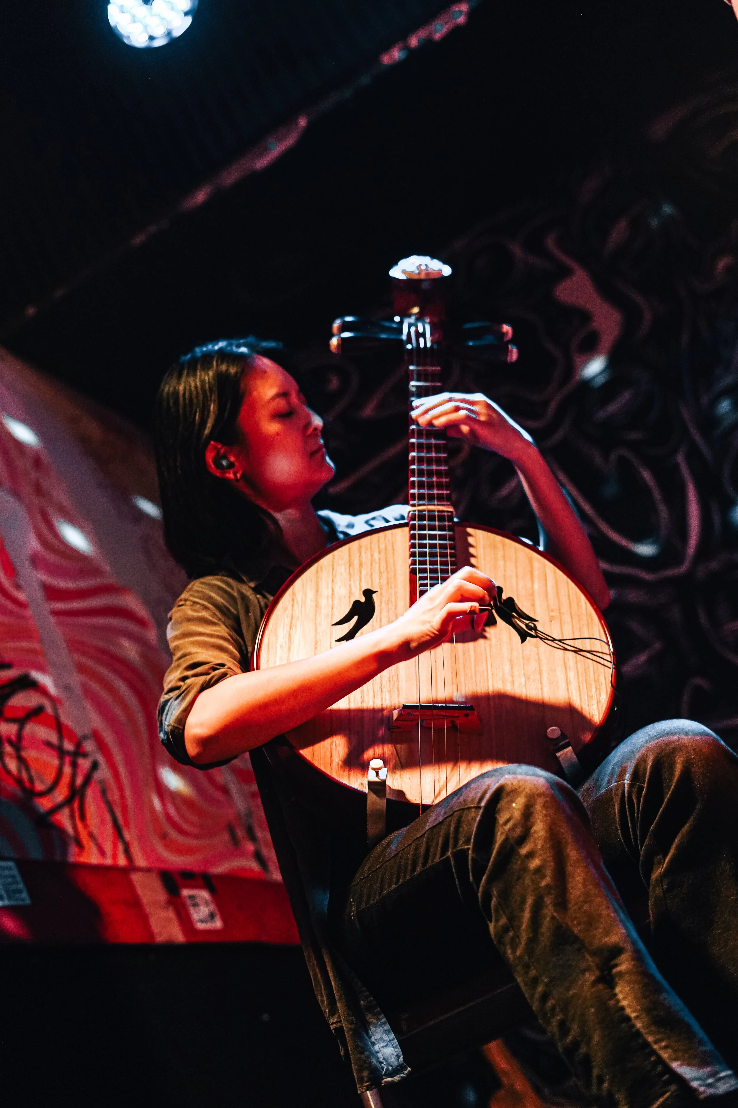 A woman playing an acoustic Chinese Ruan/ Pipa on stage at Bug Jar, Rochester, NY, with colorful abstract art in the background.
