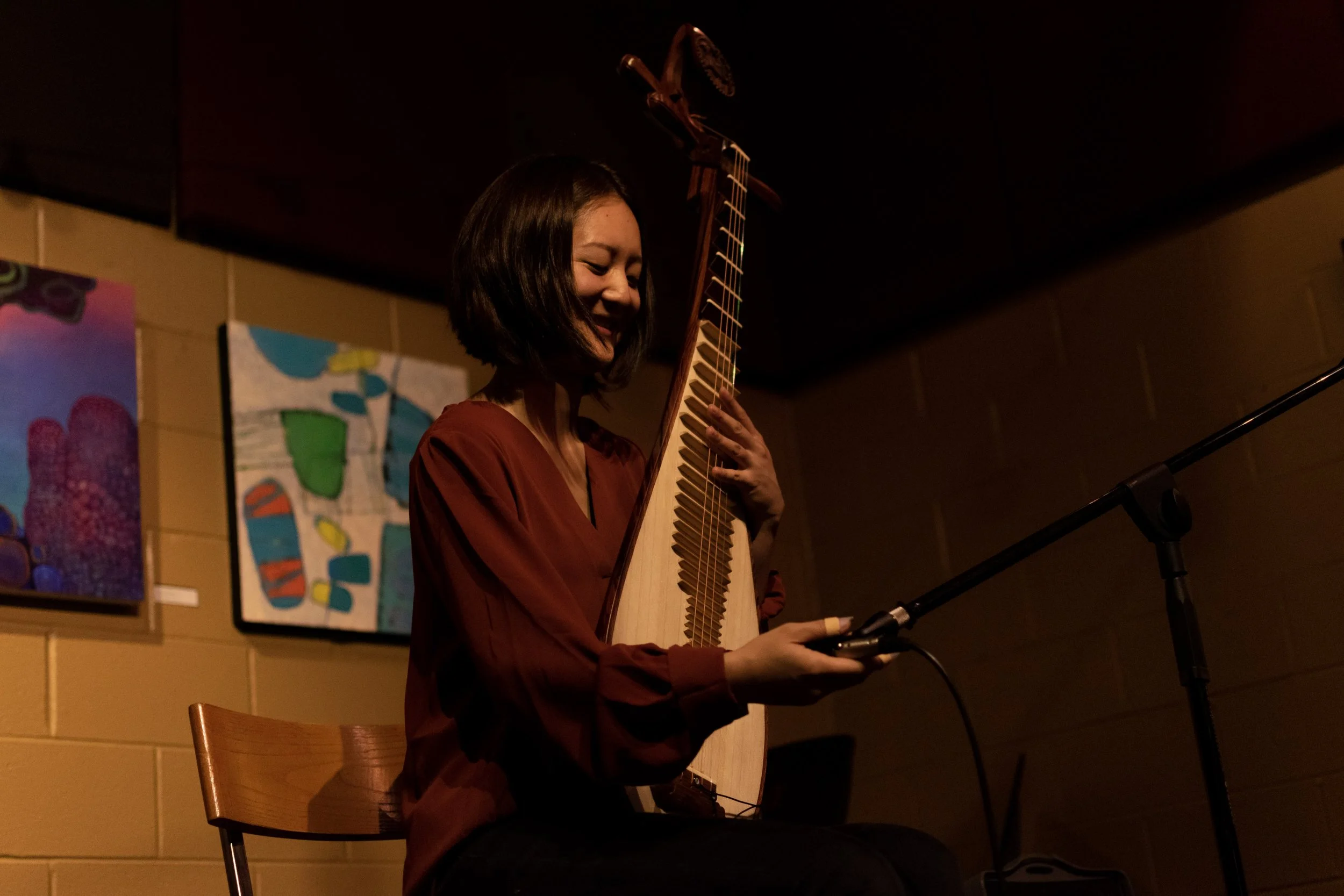 A woman with shoulder-length dark hair, at The Little Cafe, sits on a wooden chair on stage, playing an Asian/ Chinese traditional stringed instrument Chinese pipa and smiling. Behind her are colorful abstract paintings on a yellow brick wall. 