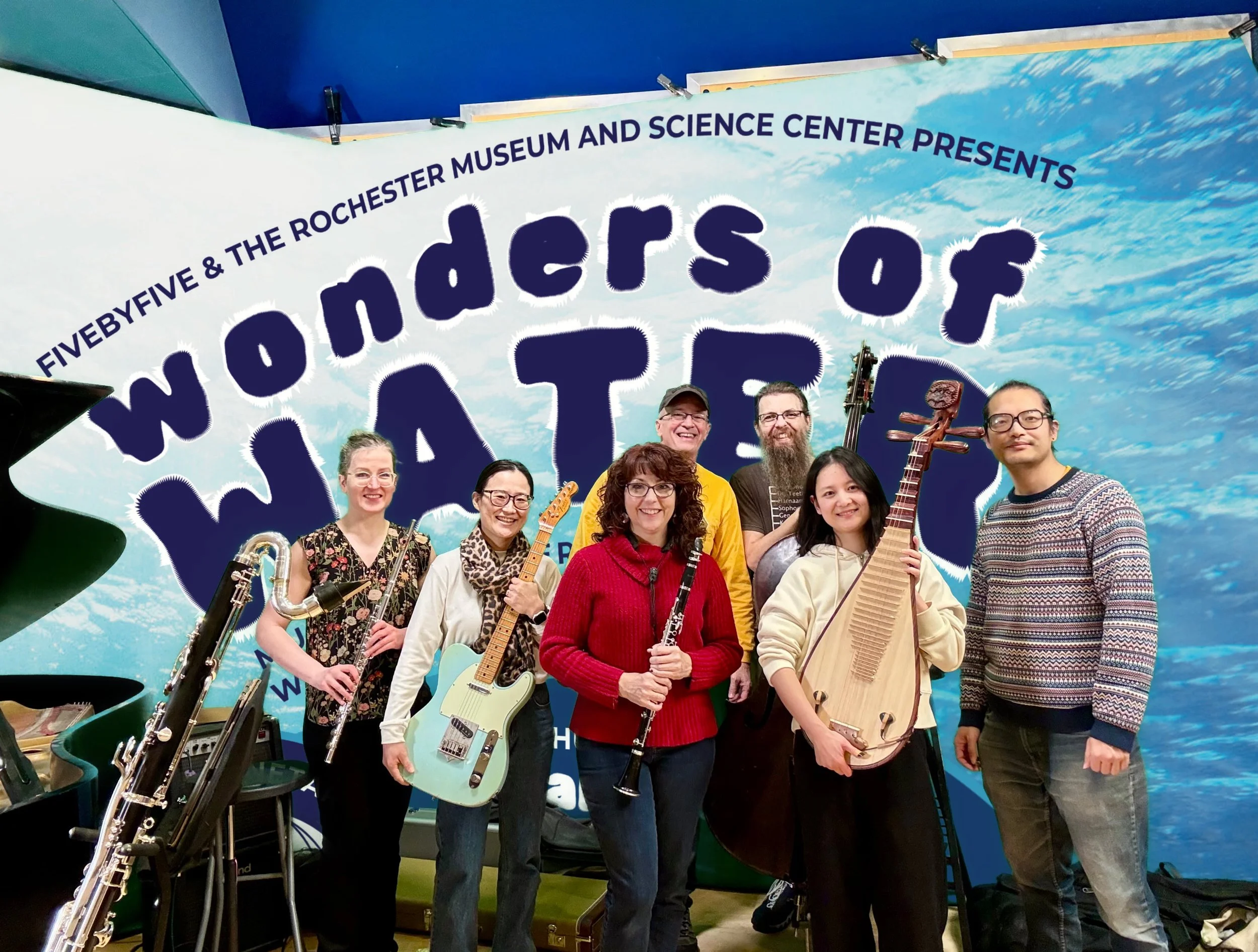 Group of seven musicians standing in front of a colorful backdrop with text that reads "Wonders of Water" and musical instruments including guitars, a clarinet, a bass, and a Chinese Pipa/ Chinese lute.
