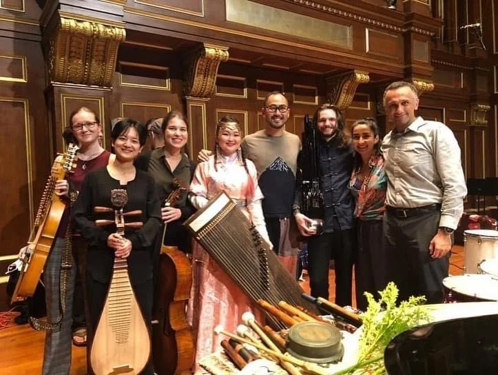 Group of nine musicians and their conductor posing in a concert hall with wood-paneled walls, holding various string instruments, including Chinese pipa and Leah Ou(O's Pipa)