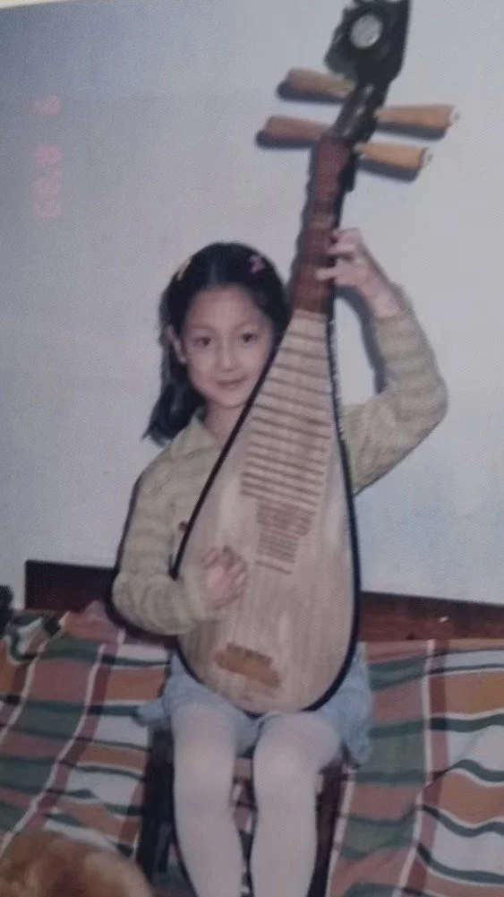 A young girl Leah Ou  sitting on a bed holding up a large wooden stringed musical instrument: Chinese pipa/ Chinese lute, smiling at the camera.