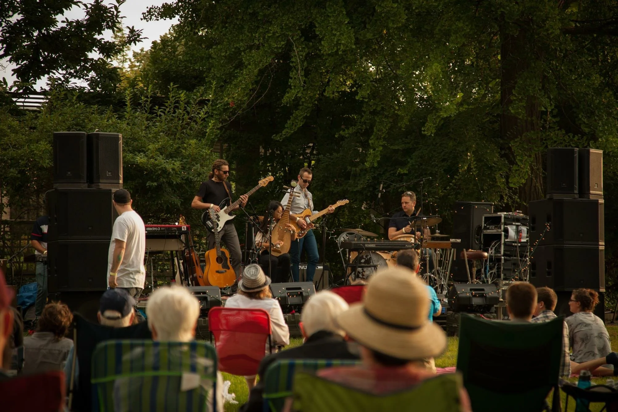 A live outdoor band with Pipa, guitar, bass and drums  performs on a stage with trees in the background in George Eastman House Concert in Rochester, NY, while an audience seated in chairs and blankets watch the performance.
