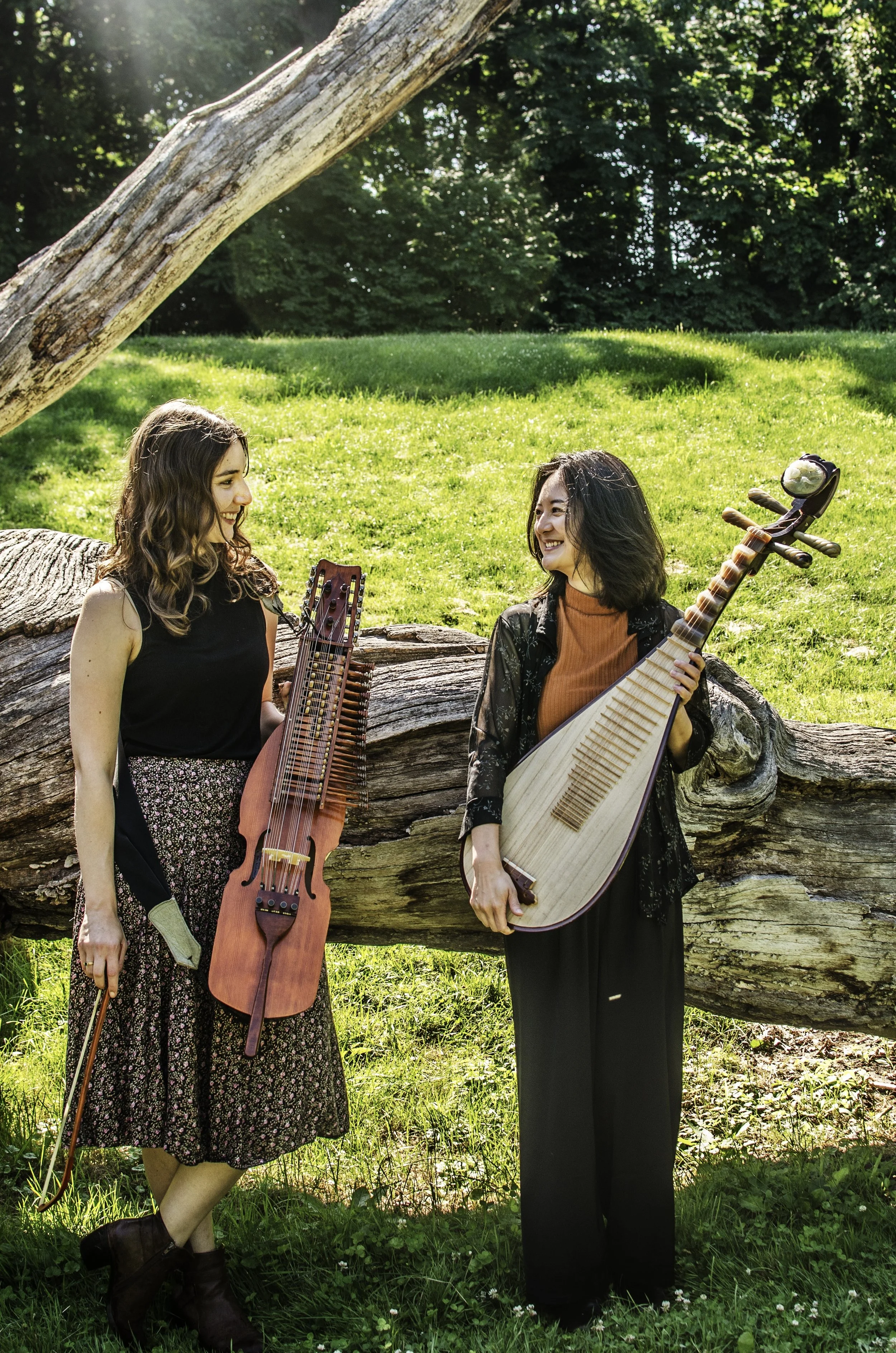 Two women standing near a fallen tree in a grassy park, smiling at each other, holding traditional stringed instruments: Chinese Lute/ Pipa. and a nyckelharpa. 