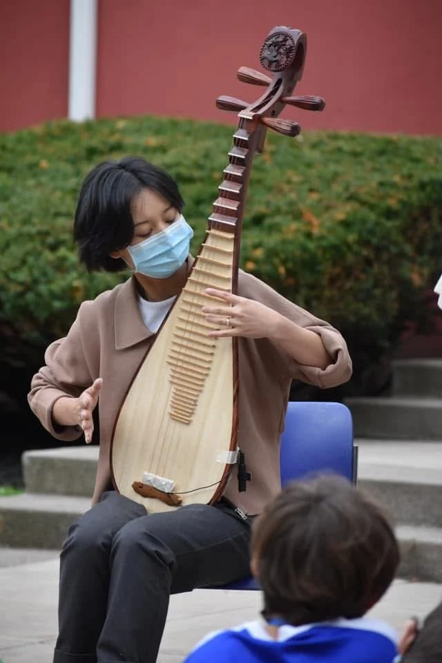 Woman wearing a face mask during Covid, at a school in Rochester, NY,  playing a traditional stringed instrument: Chinese Pipa/ Chinese Lute outdoors while students listens.