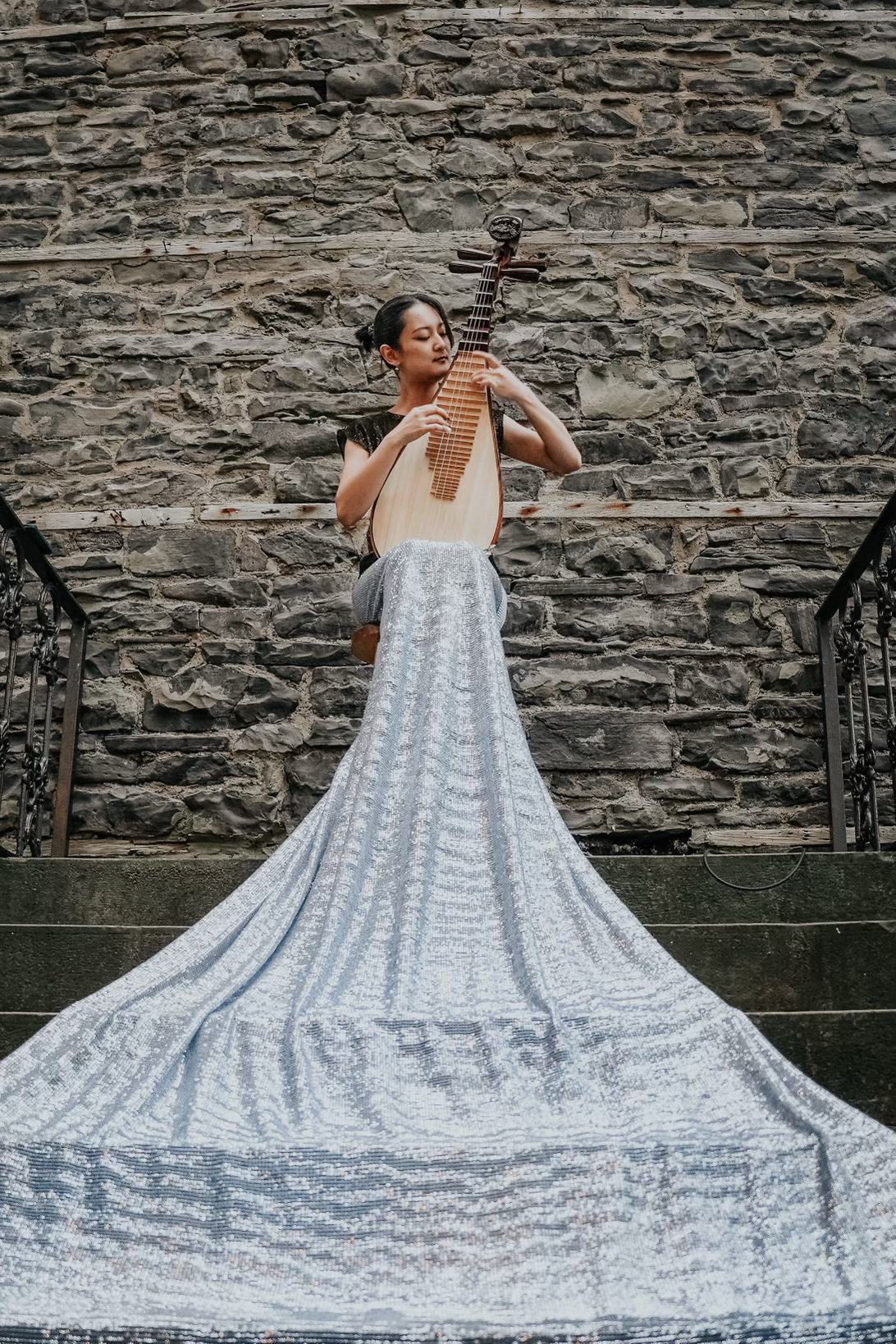 A woman sitting on a stool against a stone wall, playing a traditional stringed musical instrument: Asian pipa/ Asian Lute with a long, flowing metallic silver skirt.
