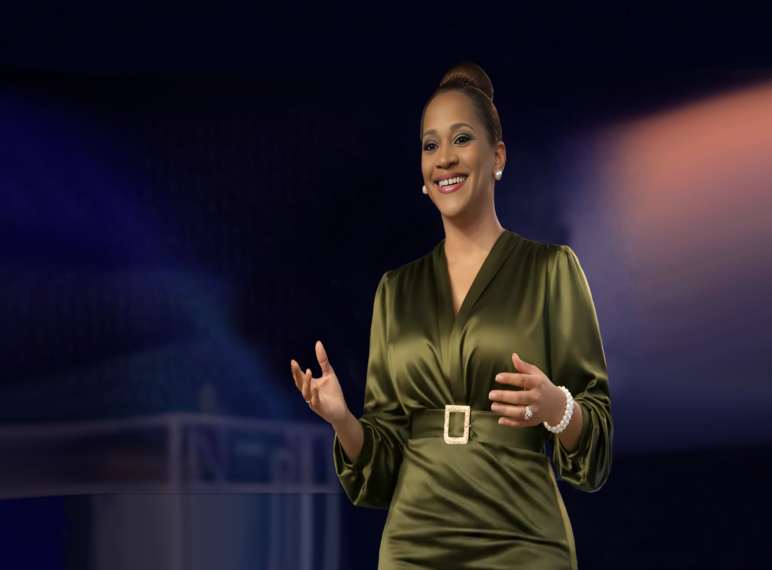 A woman in a dark green dress with a belt and pearl jewelry stands on a stage, smiling and speaking in front of a dark background with soft, colorful lighting.