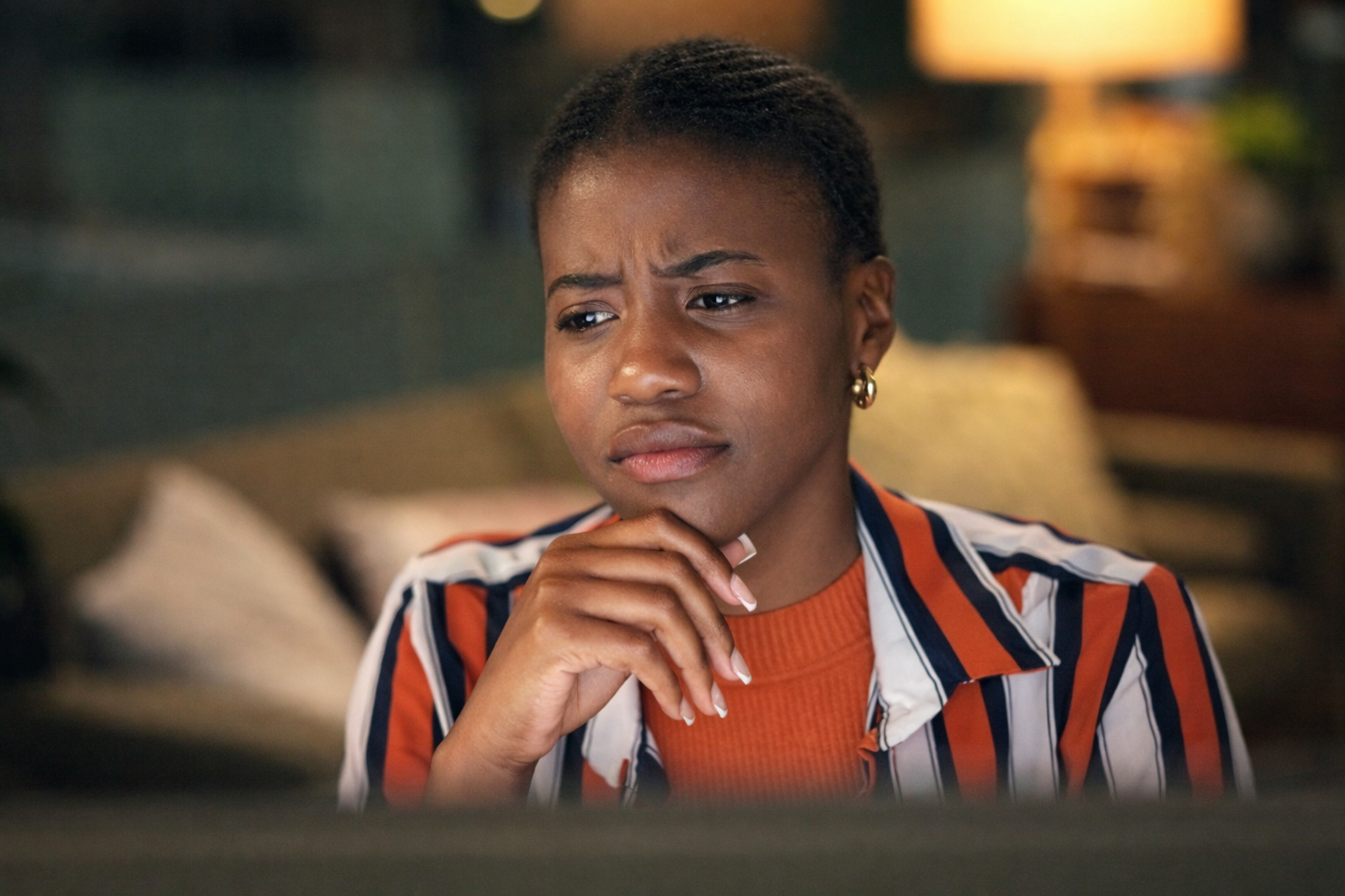 A young woman with short hair and earrings looks confused or thoughtful while sitting on a couch in a cozy, warmly lit room.