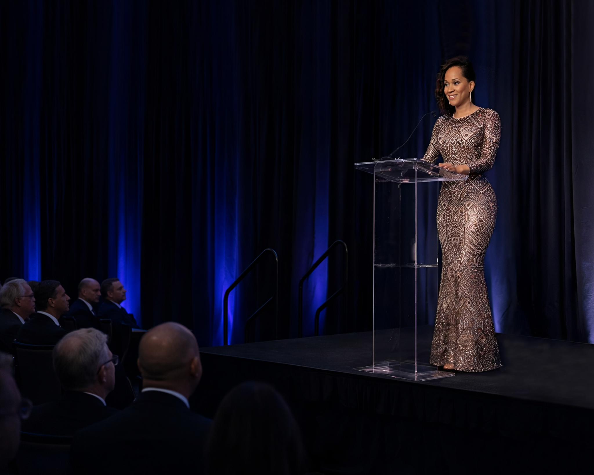A woman with dark, curly shoulder-length hair in a patterned, long-sleeve dress stands behind a clear podium, speaking into a microphone at a formal event with a dark blue curtain backdrop.