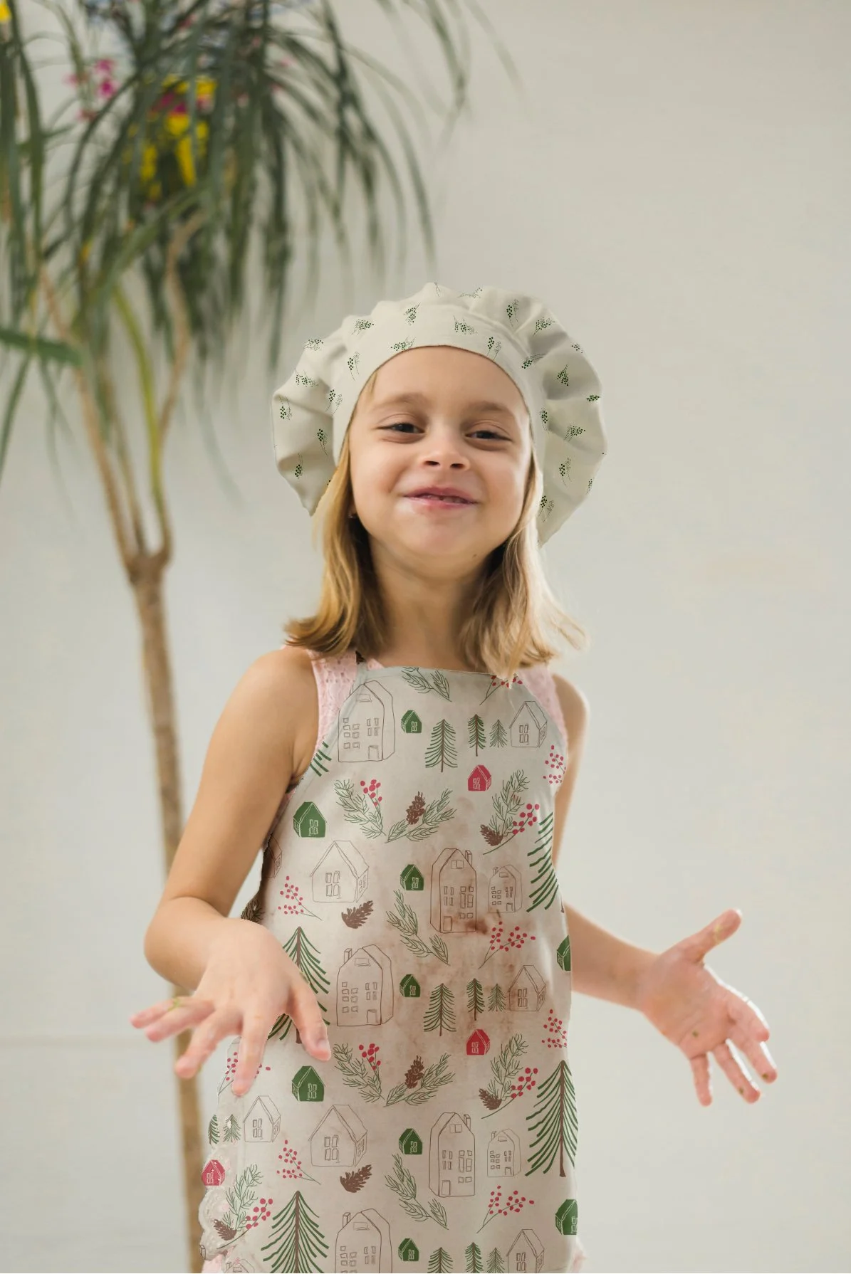 A young girl wearing a kitchen apron with a holiday pattern and a chef's hat, standing indoors near a green plant.