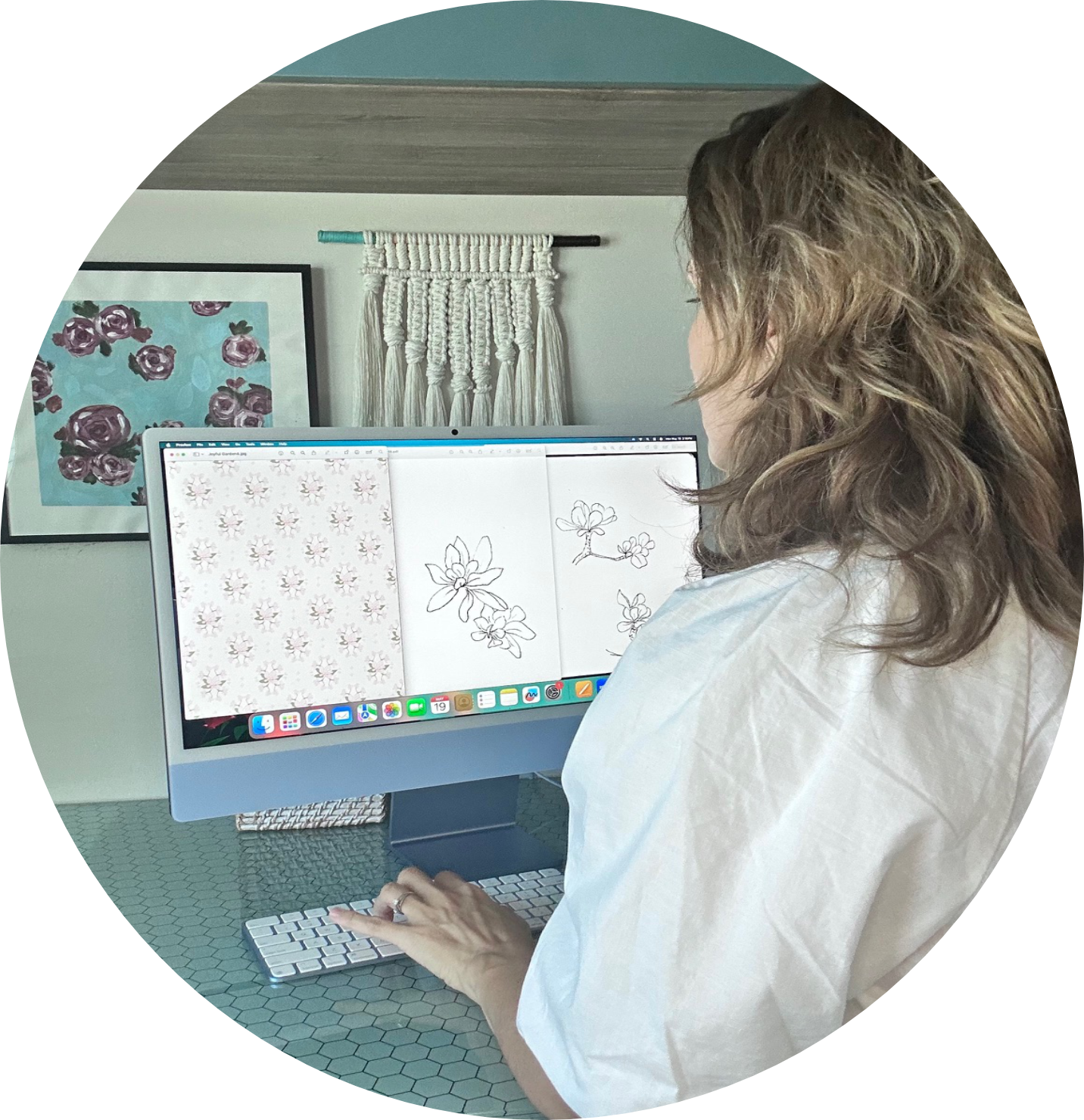 A woman with wavy, shoulder-length hair working at a desktop computer with floral sketches on the screen. She is standing at a hexagon-tiled table in a room with wall art and a macramé wall hanging. Pattern design for licensing .