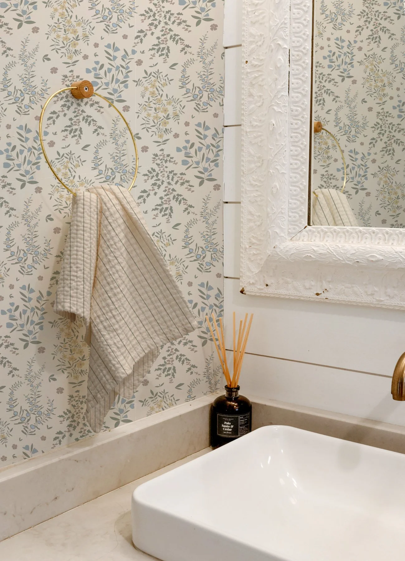 Close-up of a decorative bathroom countertop with a white oval vessel sink, a black reed diffuser labeled 'Palo Santo & Cedar,' a wall-mounted mirror with an ornate white border, a wall hook with a striped beige and white towel, and wallpaper with a floral pattern in soft muted colors.
