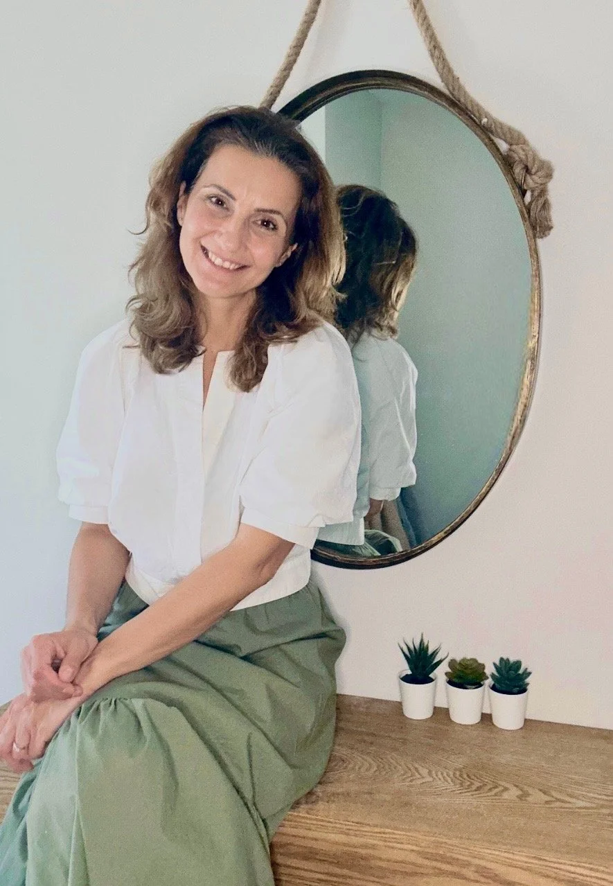 A woman with wavy brown hair smiling, sitting on a wooden surface in front of a round mirror mounted on a wall with a rope, three small potted succulents are on the surface beside her.